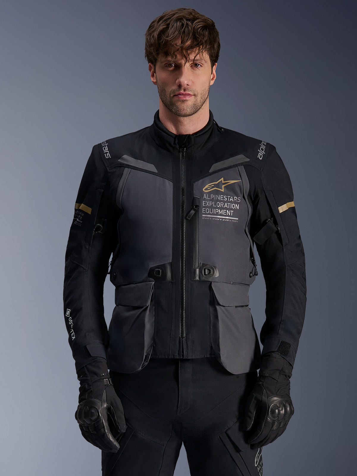 Veste Alpinestars ST-7 2L GORE-TEX, veste de moto Adventure Touring pour homme, gris foncé et noir avec accents kaki, logo « Alpinestars Exploration Equipment », poches cargo imperméables et col montant, portée par un mannequin avec des gants noirs