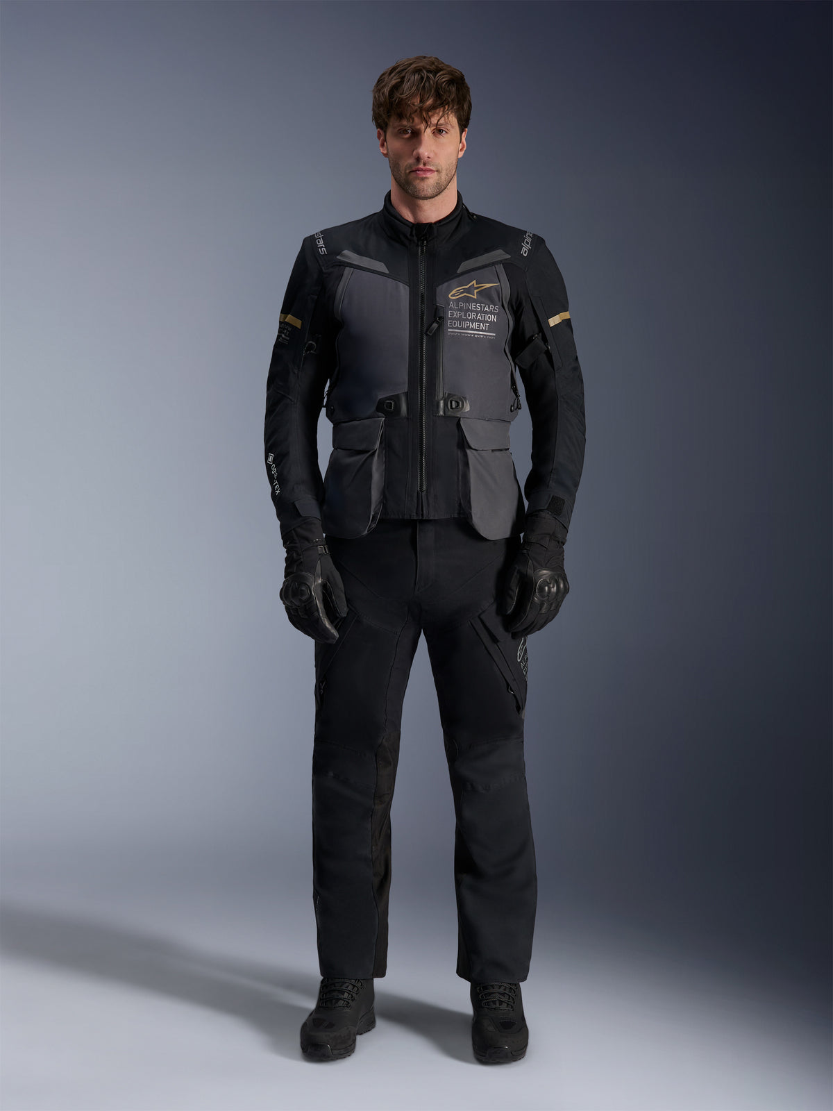 Mannequin homme portant la veste Alpinestars ST-7 2L GORE-TEX en Deep Gray Black Dark Khaki, arborant le marquage 'ALPINESTARS EXPLORATION EQUIPMENT' sur la poitrine, assortie d'un pantalon de moto, de gants et de bottes noirs, vue de face en studio.