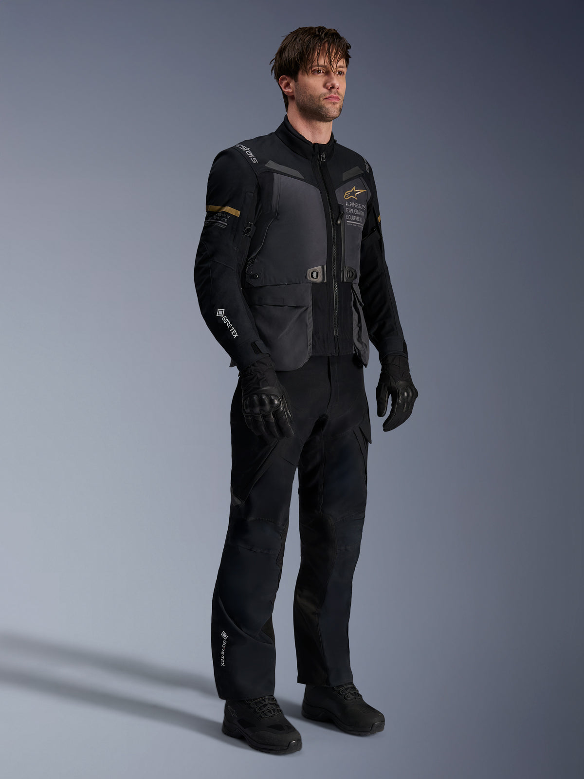 Mannequin homme portant la veste Alpinestars ST-7 2L GORE-TEX en Deep Gray Black et Dark Khaki, associée à un pantalon d'aventure et des gants noirs assortis, avec une coque laminée imperméable, des poches cargo et un marquage technique, idéale pour le touring d'aventure.