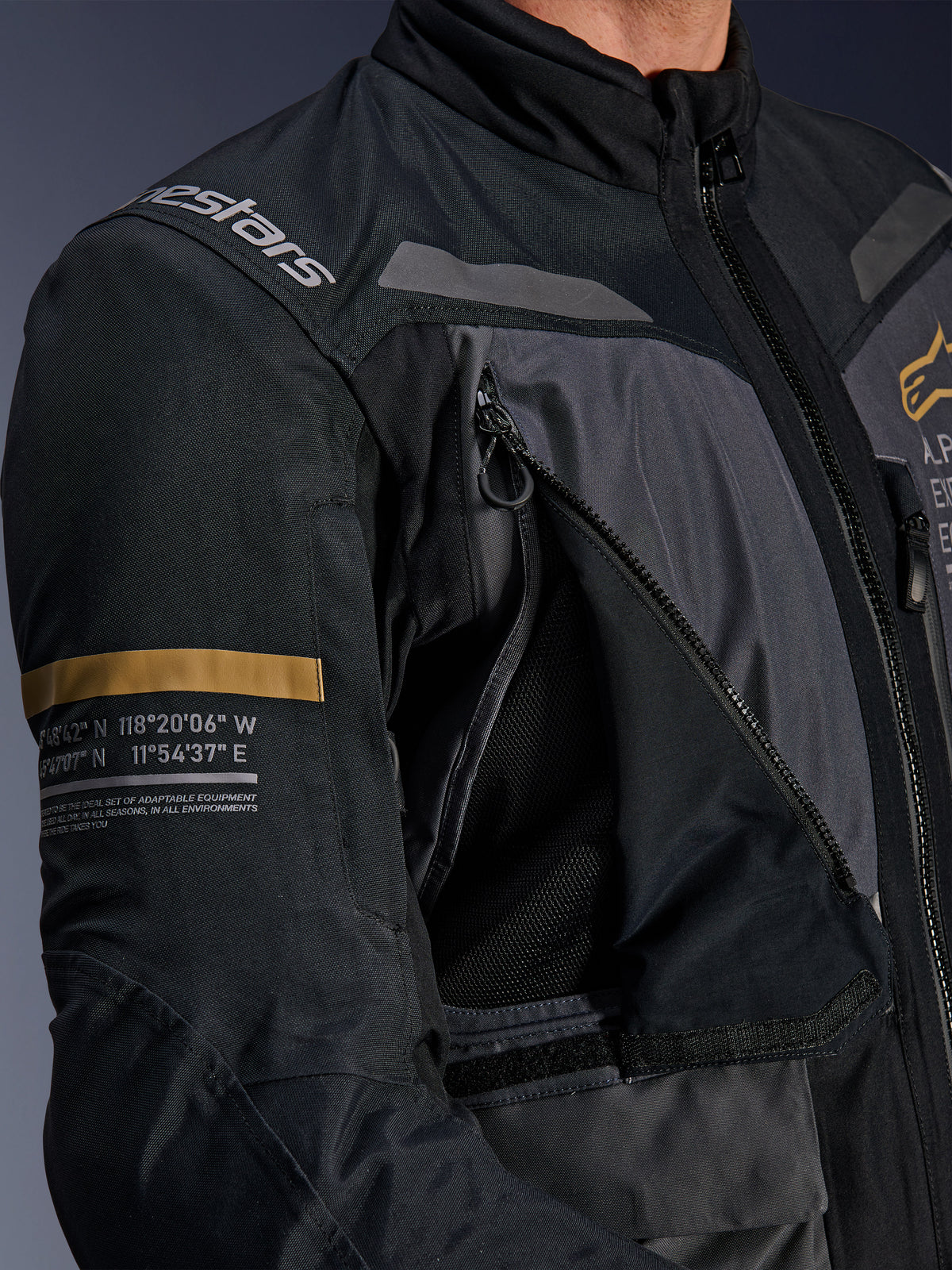 Veste Alpinestars ST-7 2L GORE-TEX, gris foncé et noir, gros plan montrant le panneau de ventilation zippé ouvert sur la poitrine avec maille, logo Alpinestars sur l'épaule et bande kaki sur la manche avec coordonnées géographiques imprimées, équipement de moto technique adventure-touring