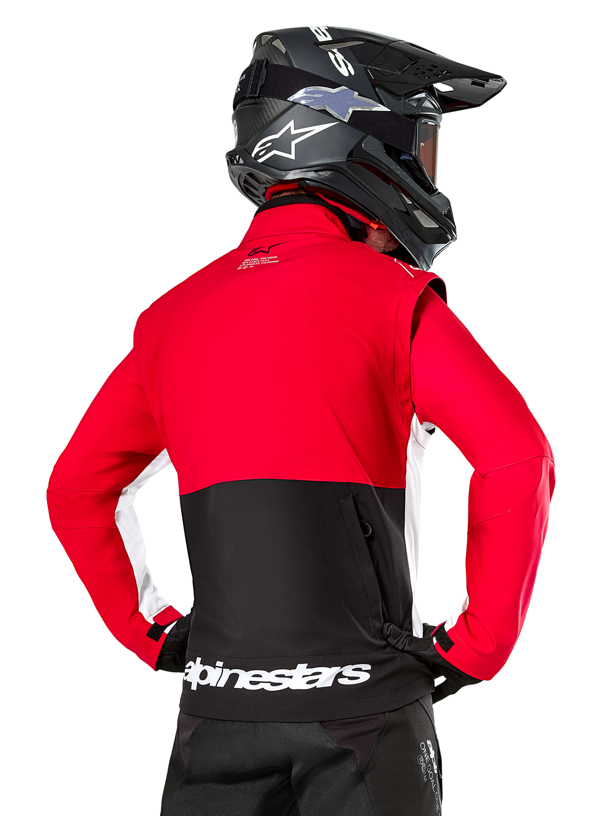 Veste Softshell Alpinestars Lite-Dura, Noir Rouge Vif Blanc, vue arrière d'un pilote de motocross, haut du corps et manches rouges avec empiècement noir en bas du dos, poche de rangement arrière zippée, logo Alpinestars blanc sur l'ourlet inférieur, portée avec un casque et un pantalon noirs