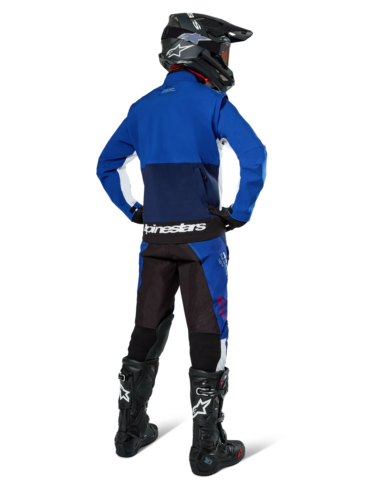 Pilote de motocross vu de dos portant la veste softshell Alpinestars Lite-Dura en bleu marine et blanc, avec un pantalon d'enduro assorti, un casque noir et des bottes noires, équipement de course tout-terrain complet