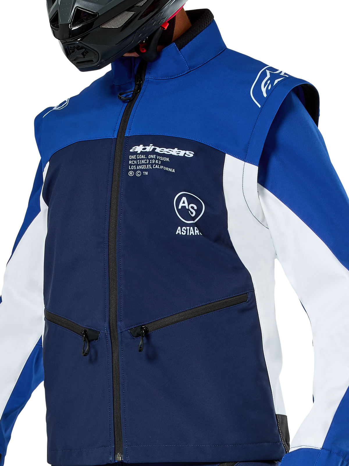 Veste Softshell Alpinestars Lite-Dura, veste de motocross et d'enduro, bleu marine, bleu vif et blanc, avec logo Alpinestars blanc sur la poitrine et poches zippées, portée avec un casque noir, construction softshell durable avec manches amovibles pour la conduite tout-terrain.