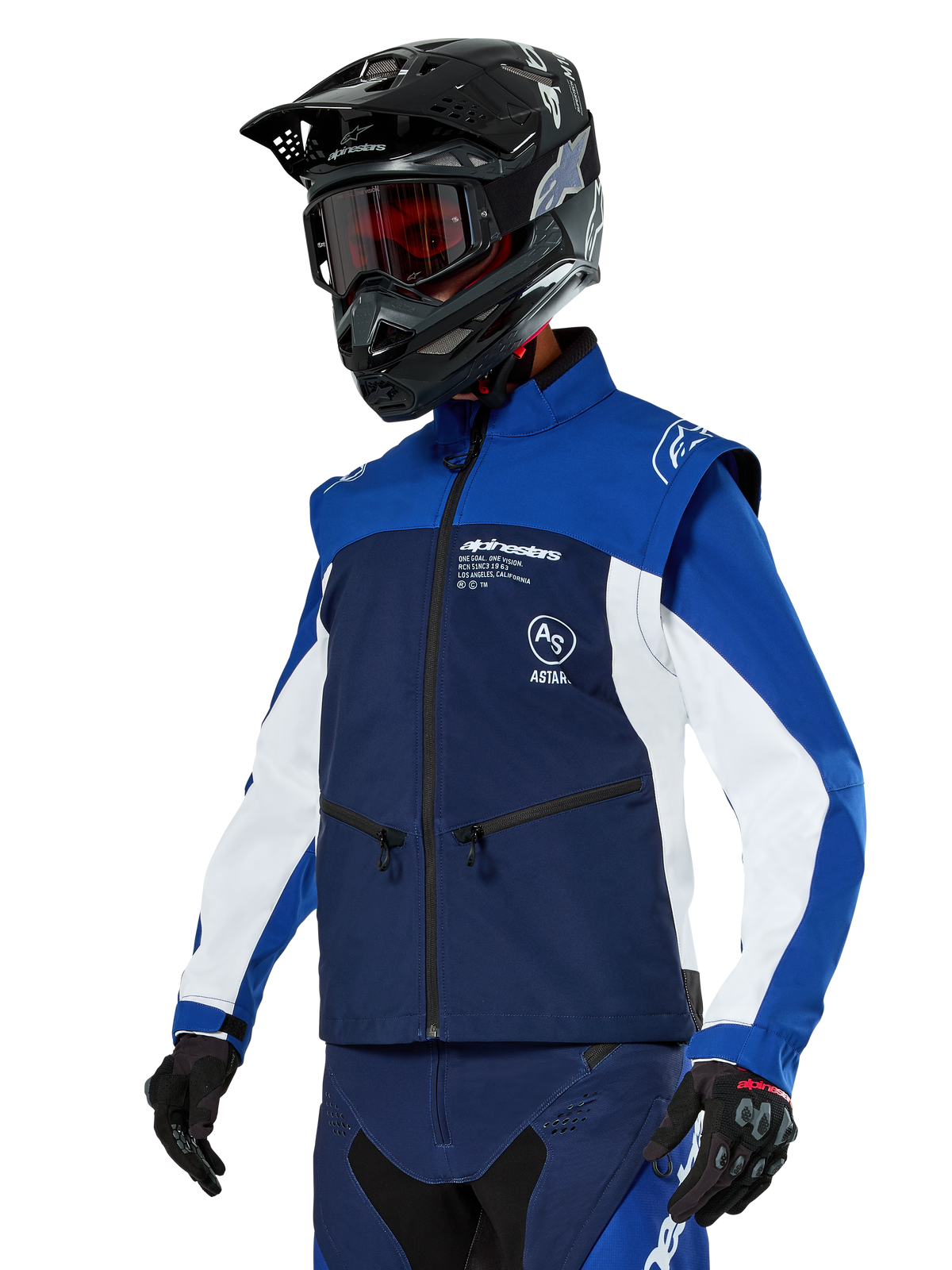 Veste softshell Alpinestars Lite-Dura, bleu marine, bleu vif et blanc, veste d'enduro tout-terrain, portée avec un casque noir et des lunettes teintées rouges, design color-block sportif avec plusieurs fermetures éclair et logos de la marque, textile résistant pour le motocross et le trail