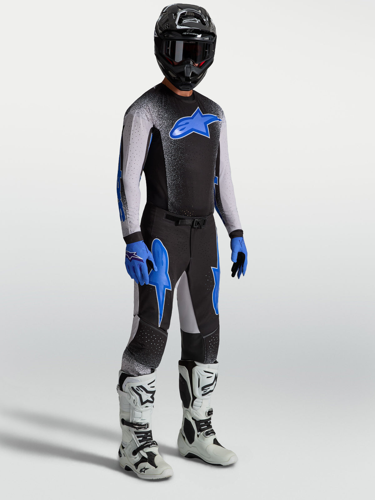 Pantalons Supertech Hiten AFD Alpinestars, pantalons de motocross pour homme, noirs et bleus, coupe athlétique performance, portés sur mannequin avec maillot assorti et bottes de moto blanches.