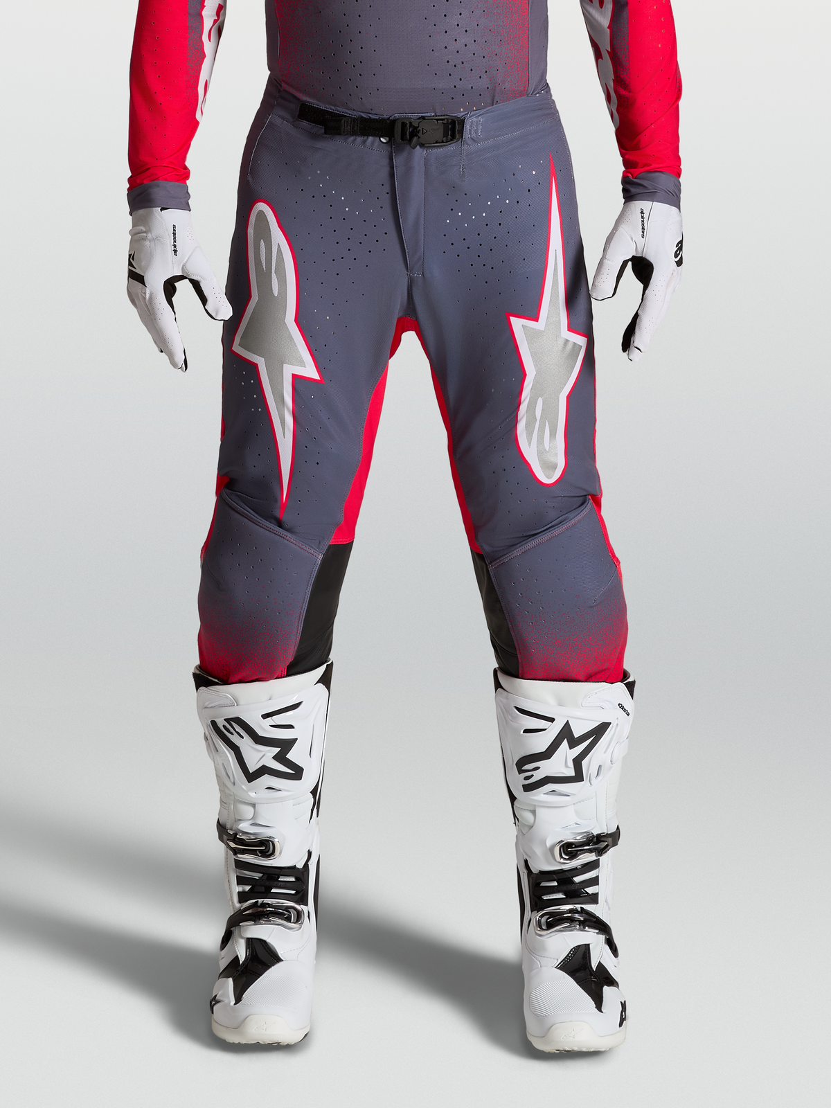 Alpinestars Supertech Hiten AFD, pantalon de motocross haute performance, gris et rouge fluo avec graphismes étoilés lilas clair, empiècements stretch perforés, porté avec des bottes blanches