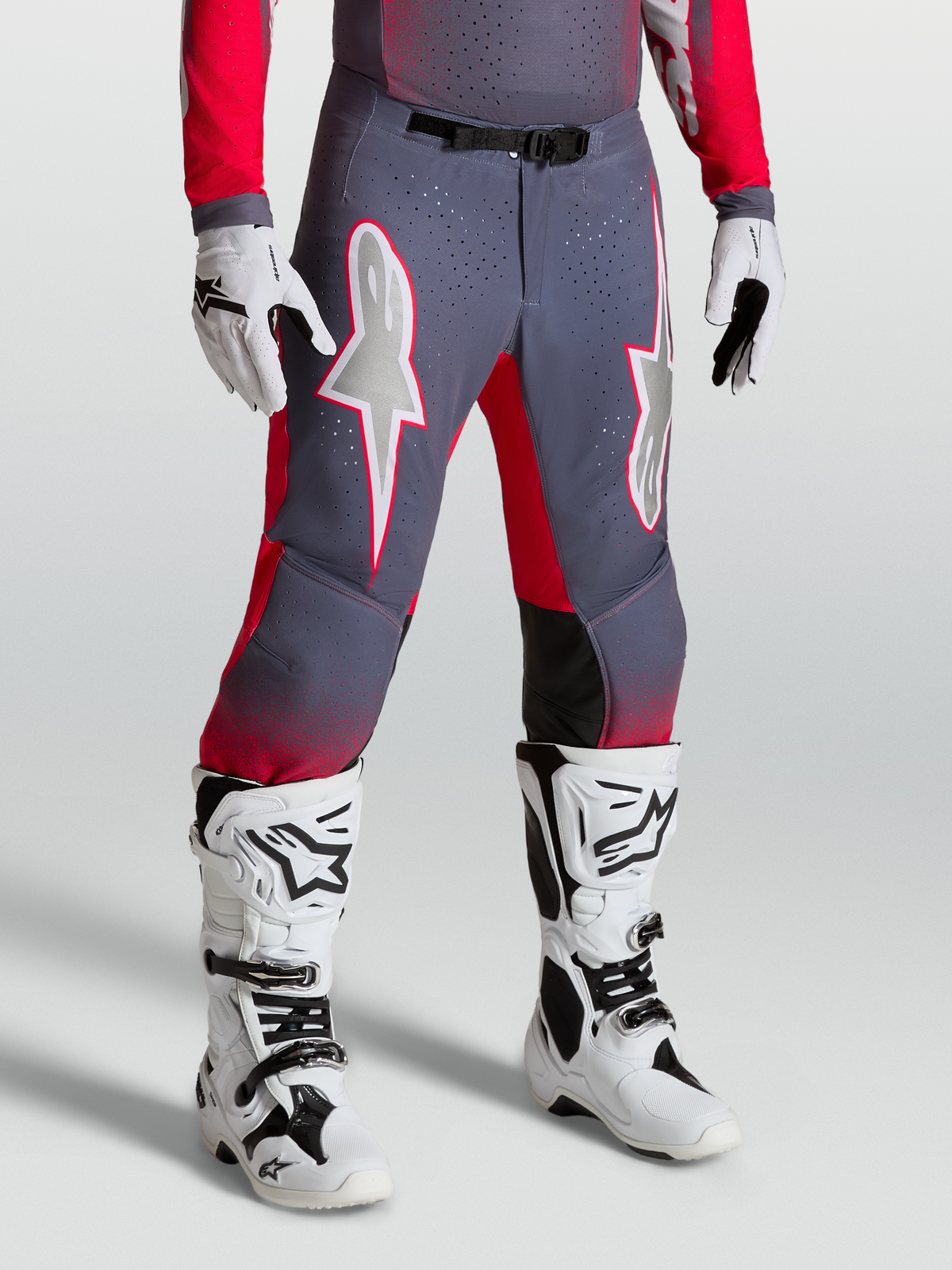 Alpinestars Supertech Hiten AFD, pantalon de motocross gris et rouge fluo avec graphismes d'étoiles blanches et boucle magnétique, porté avec des bottes blanches