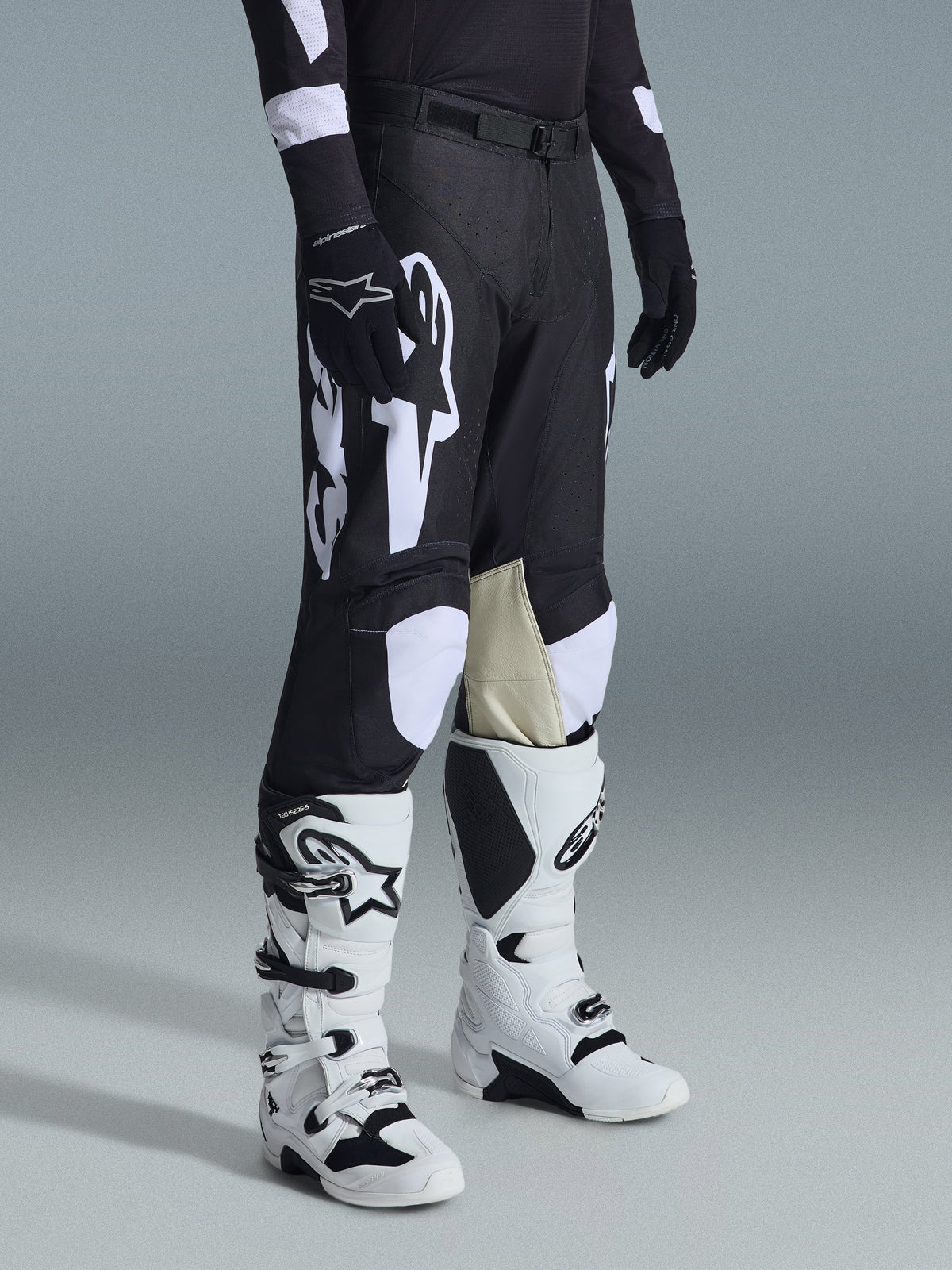 Pantalon Alpinestars 2026 Racer Air Riway, pantalon de motocross noir et blanc avec de grands graphismes de logo blancs, empiècements en cuir beige à l'intérieur du genou, porté avec des bottes tout-terrain blanches et noires et des gants noirs dans un décor de studio.