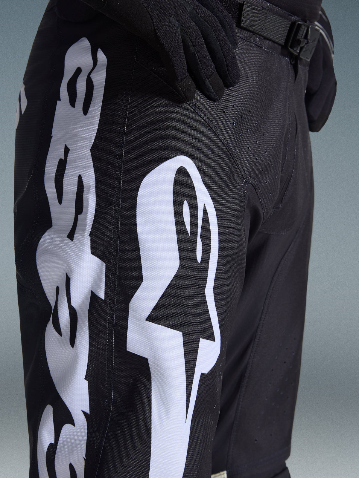 Pantalon Alpinestars Racer Air Riway 2026, pantalon de motocross noir avec un logo Alpinestars blanc sur la cuisse, gros plan montrant le tissu technique et la sangle de réglage à la taille, porté avec des gants noirs pour le pilotage tout-terrain