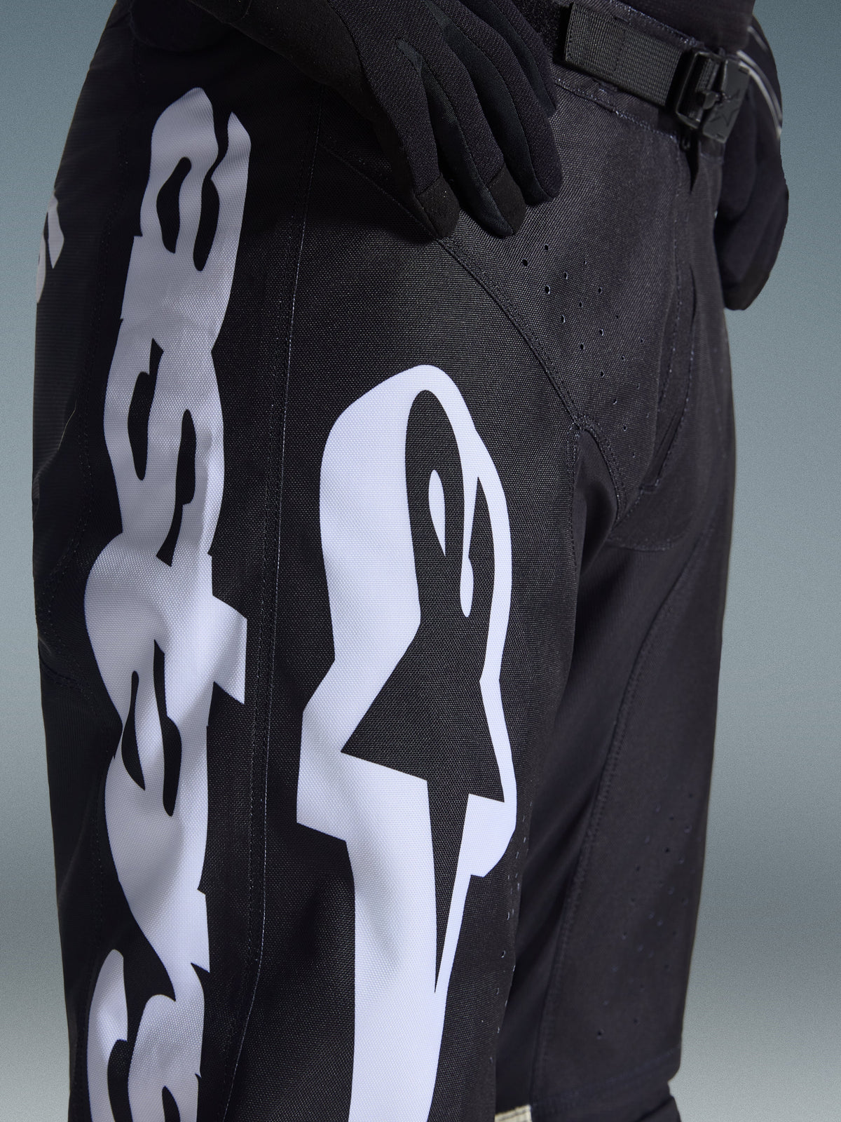 Alpinestars 2026 Racer Air Riway Pants, pantalon de motocross, noir et blanc, vue rapprochée montrant le grand logo Alpinestars blanc, les perforations découpées au laser pour la circulation de l'air et la boucle de taille réglable, tissu durable pour la conduite tout-terrain.
