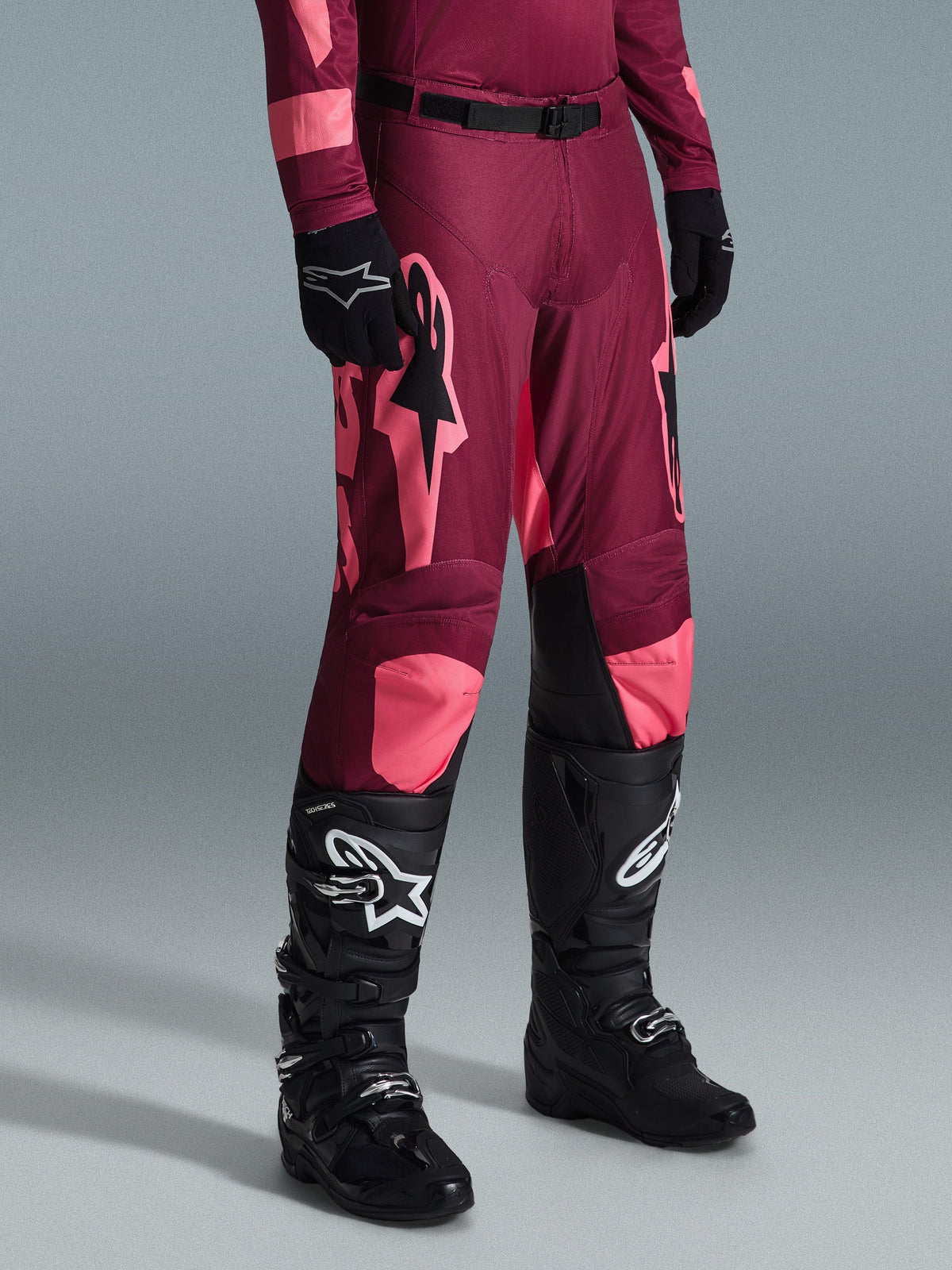 Alpinestars 2026 Racer Riway Pants, pantalon de motocross, rouge corail foncé, avec de grands logos étoiles roses et des panneaux intérieurs noirs aux genoux, porté avec des bottes de motocross noires et des gants noirs, coupe de course athlétique pour le pilotage tout-terrain