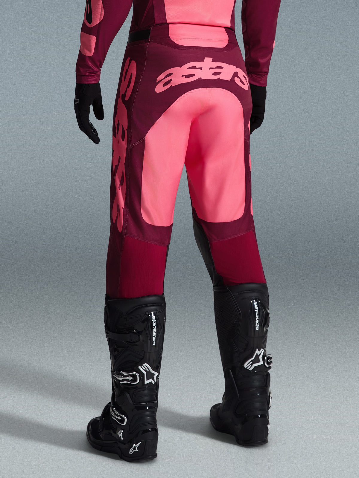 Pantalon Alpinestars Racer Riway 2026, pantalon de motocross, rouge foncé et corail, vue arrière montrant le grand logo 'astars' corail sur l'assise, porté avec des bottes et des gants de motocross noirs, équipement de performance pour le tout-terrain