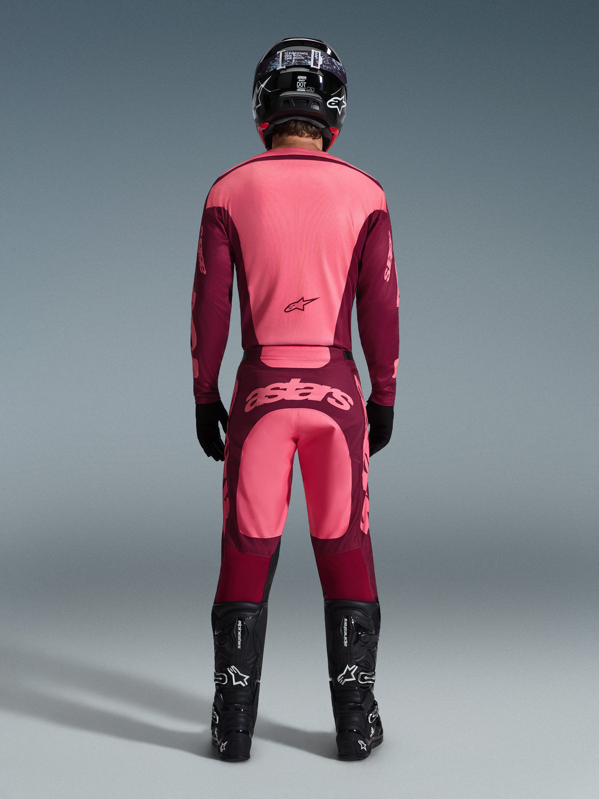 Vue arrière d'un pilote de motocross portant l'ensemble d'équipement Racer Riway 2026, maillot rose et bordeaux, pantalon bordeaux assorti avec logo « astars » rose clair sur l'assise, casque noir, bottes noires et gants noirs, tenue coordonnée pour la performance tout-terrain