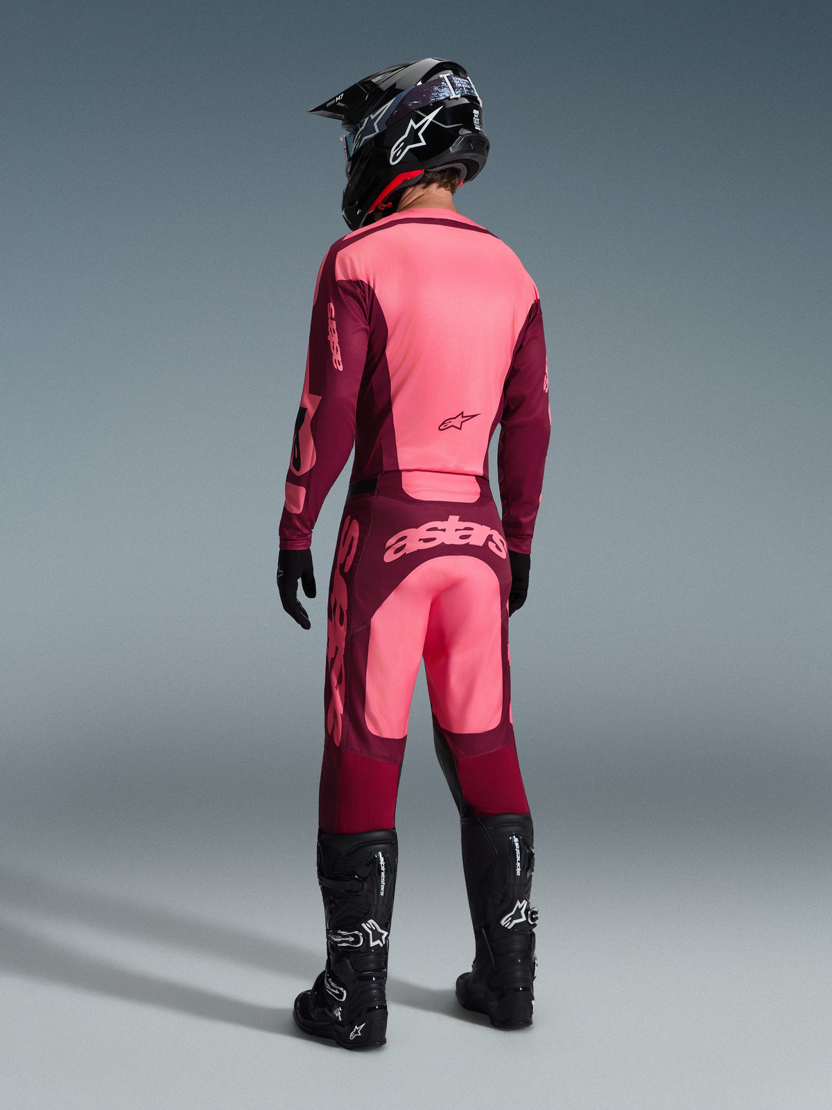 Pilote de motocross portant l'ensemble d'équipement Alpinestars Racer Riway 2026 vu de dos, coloris bordeaux et rose clair, maillot et pantalon avec logos distinctifs, complété par un casque, des gants et des bottes noirs, tenue de course de motocross coordonnée