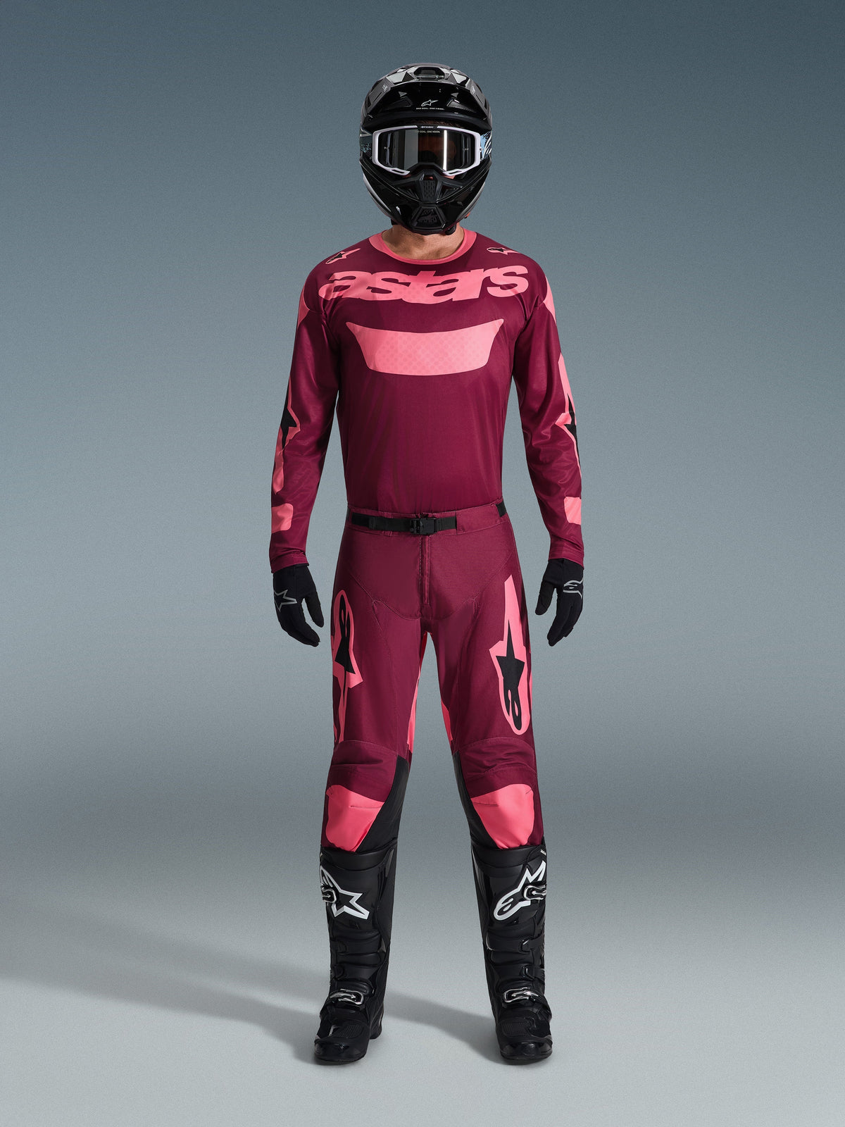 Pilote de motocross Alpinestars avec équipement de course complet, maillot Dark Red Coral avec accents roses, pantalon Racer Riway assorti, casque noir avec détails blancs, bottes noires avec logos blancs, gants noirs, tenue coordonnée pour la course tout-terrain