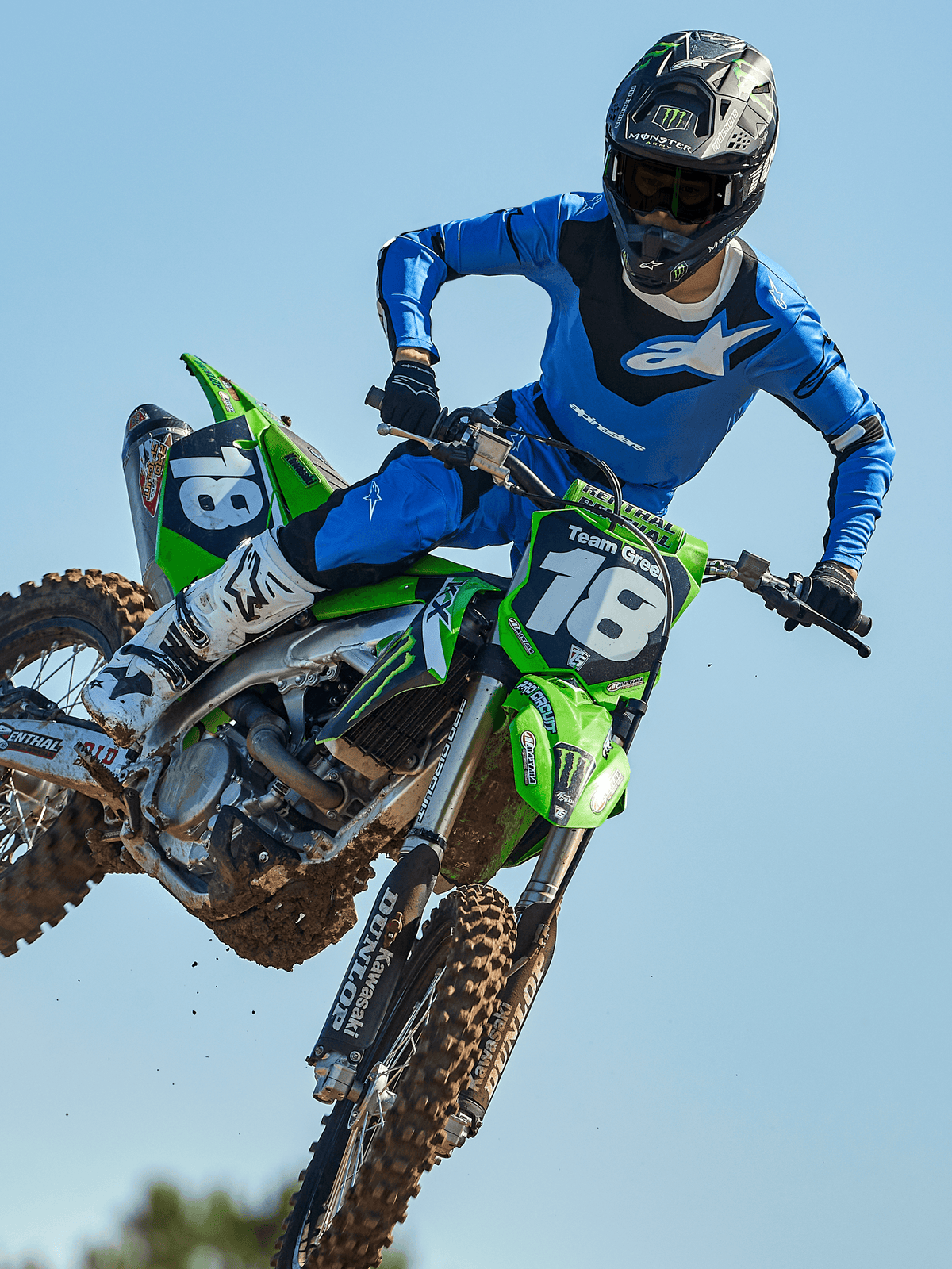 Pilote de motocross en plein saut sur une moto verte, portant le pantalon Alpinestars Racer Veil 2025 bleu et noir, un maillot bleu assorti avec logo blanc sur la poitrine, des bottes blanches et noires et un casque noir, équipement tout-terrain haute performance pour la course.