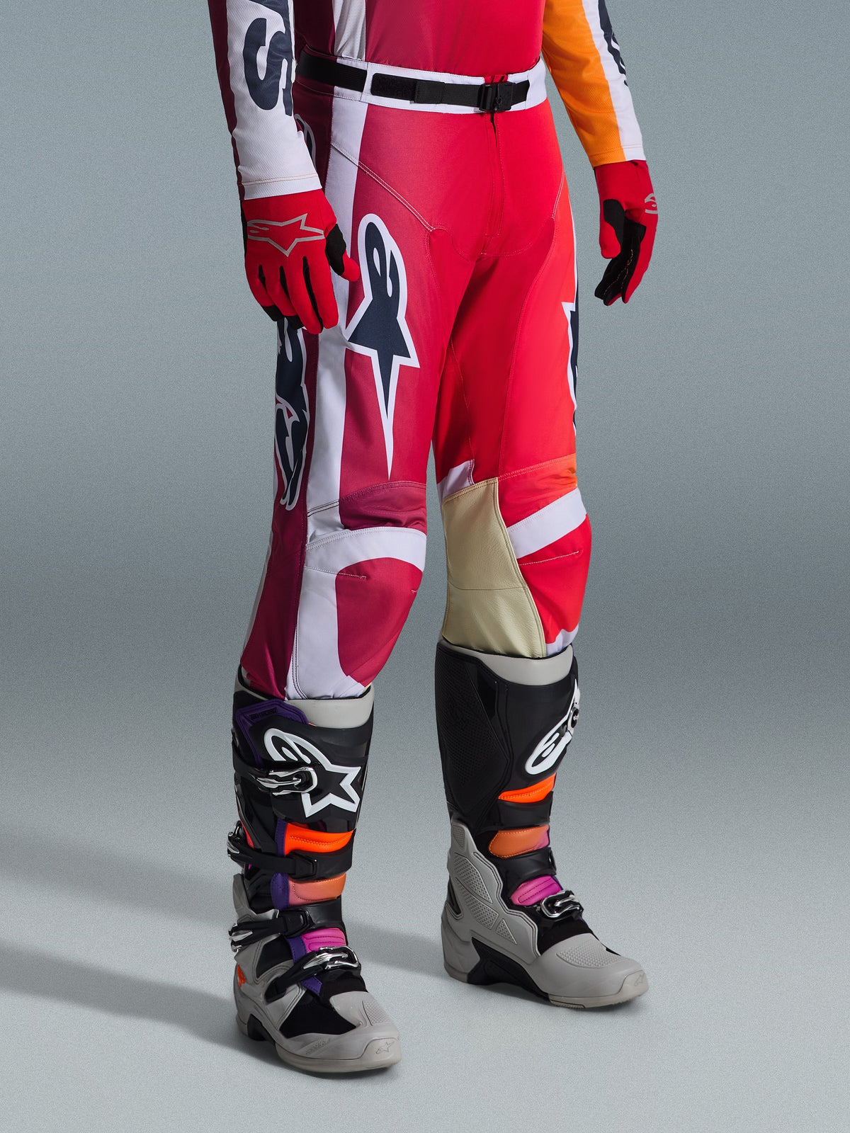 Pantalon Alpinestars 2026 Racer Portl, pantalon de motocross, rouge, gris clair et gris foncé, porté avec des gants rouges assortis et des bottes de motocross multicolores, doté de logos Alpinestars imposants et de panneaux renforcés pour la course tout-terrain.