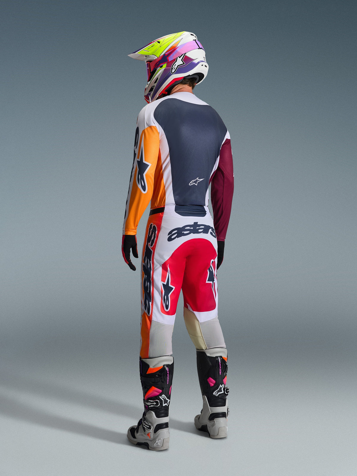 Pilote de motocross vu de dos portant une tenue de course complète, pantalon Alpinestars Racer Portl 2026 en rouge, gris clair et gris foncé, maillot assorti avec manches orange et bordeaux, casque aux graphismes multicolores, bottes tout-terrain grises et noires avec des touches de rose, tenue de motocross professionnelle