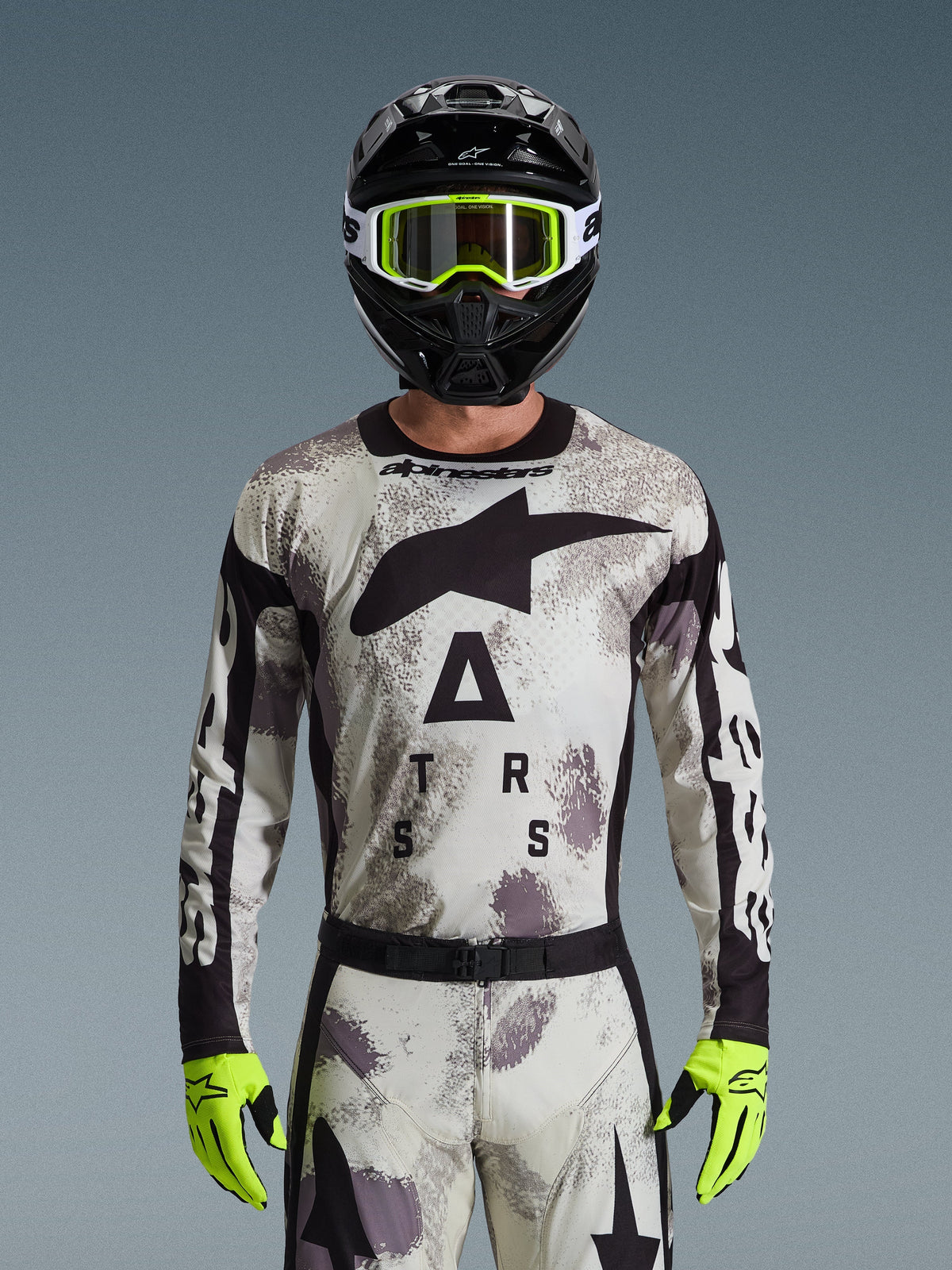 Motocross rider wearing Alpinestars 2026 Racer Lahnd Jersey, Sand Camo pattern in gray and white, large black Astar logo and branding on the chest, paired with a black helmet, neon yellow goggles and gloves, performance gear for off-road riding