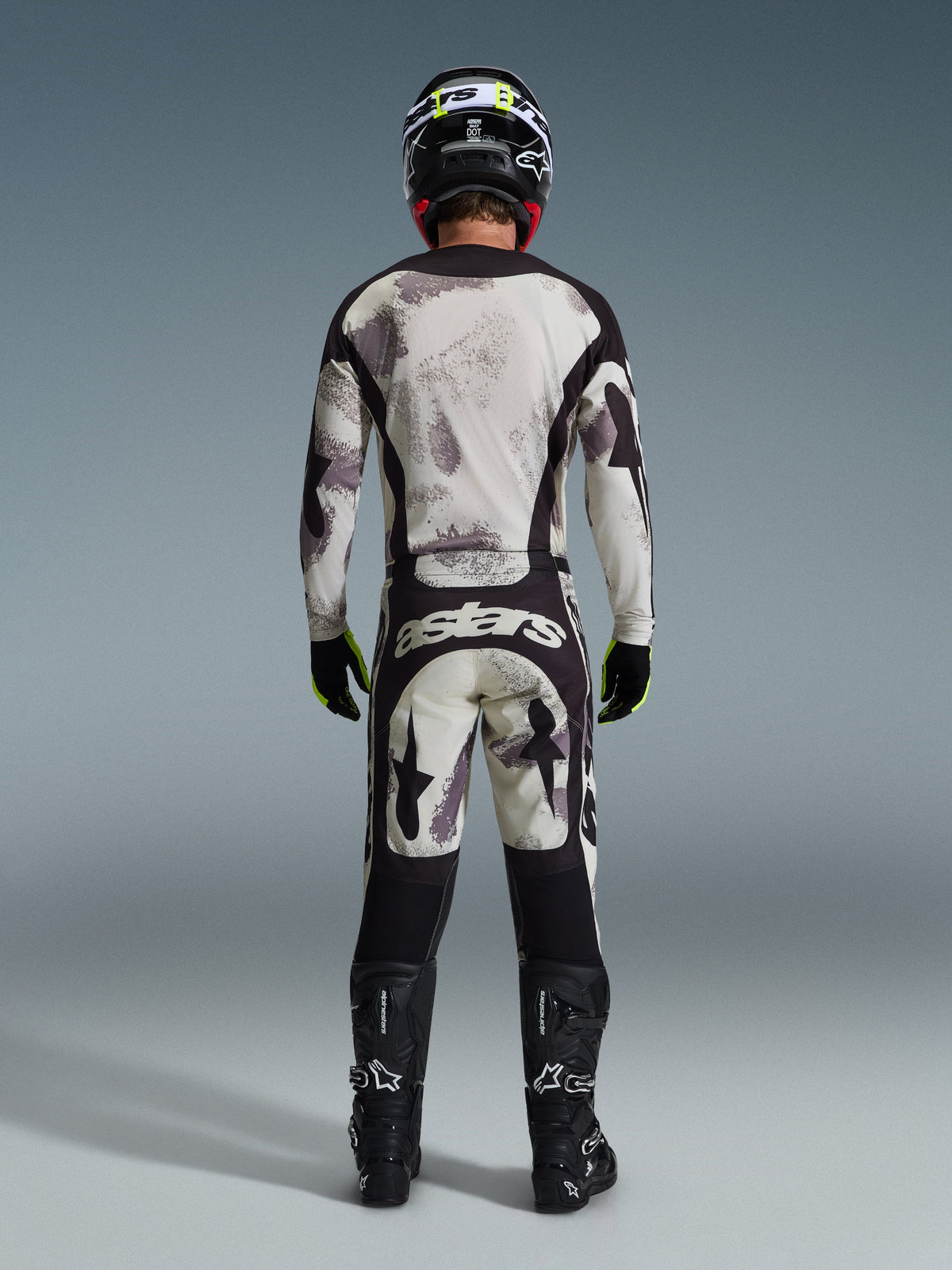 Pilote de motocross vu de dos portant le pantalon Alpinestars Racer Lahnd 2026 en Sand Camo, maillot à manches longues assorti avec motif camouflage gris, bottes de motocross noires, casque noir et marquage « astars » sur l'assise, conçu pour la course tout-terrain professionnelle.