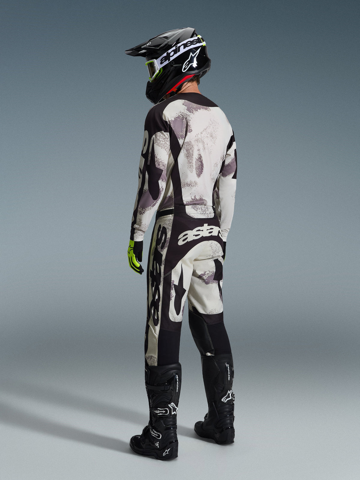 Pilote de motocross portant le pantalon Alpinestars 2026 Racer Lahnd en coloris Sand Camo et gris, tenue de course complète avec maillot assorti, casque noir et bottes noires, vue arrière mettant en valeur le marquage audacieux et le design ergonomique.