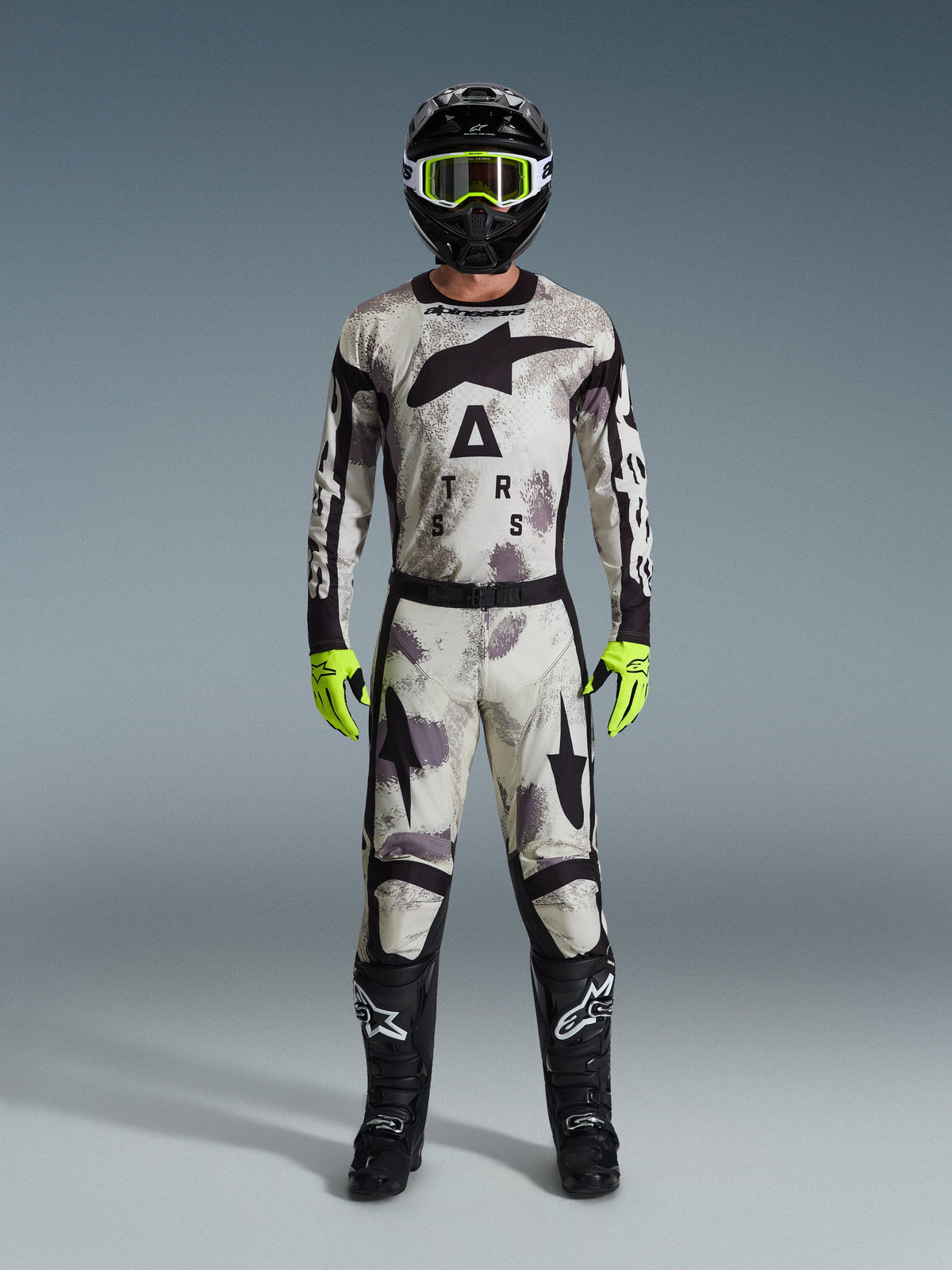 Pilote de motocross portant une tenue complète Alpinestars 2026, pantalon Racer Lahnd et maillot assorti en gris Sand Camo, casque noir avec lunettes jaune fluo, gants jaune fluo et bottes noires, tenue professionnelle coordonnée pour le tout-terrain