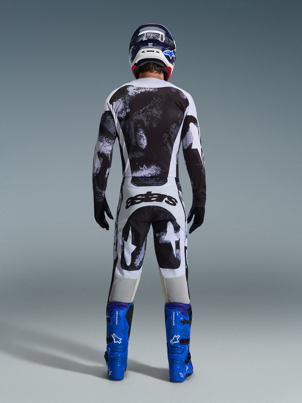 Pilote de motocross vu de derrière portant le maillot Alpinestars Racer Lahnd 2026 et le pantalon assorti en Iron Camo, avec un motif d'éclaboussures noir et blanc, bottes bleu vif et casque coordonné, tenue complète pour la course tout-terrain