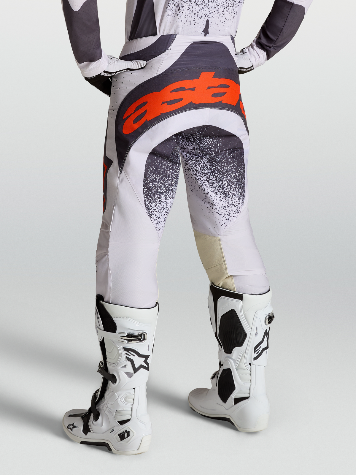 Alpinestars Techstar Swan, pantalon de motocross pour homme, lilas clair et gris foncé avec accents rouge fluo, porté avec des bottes Tech 10 blanches, idéal pour le racing off-road