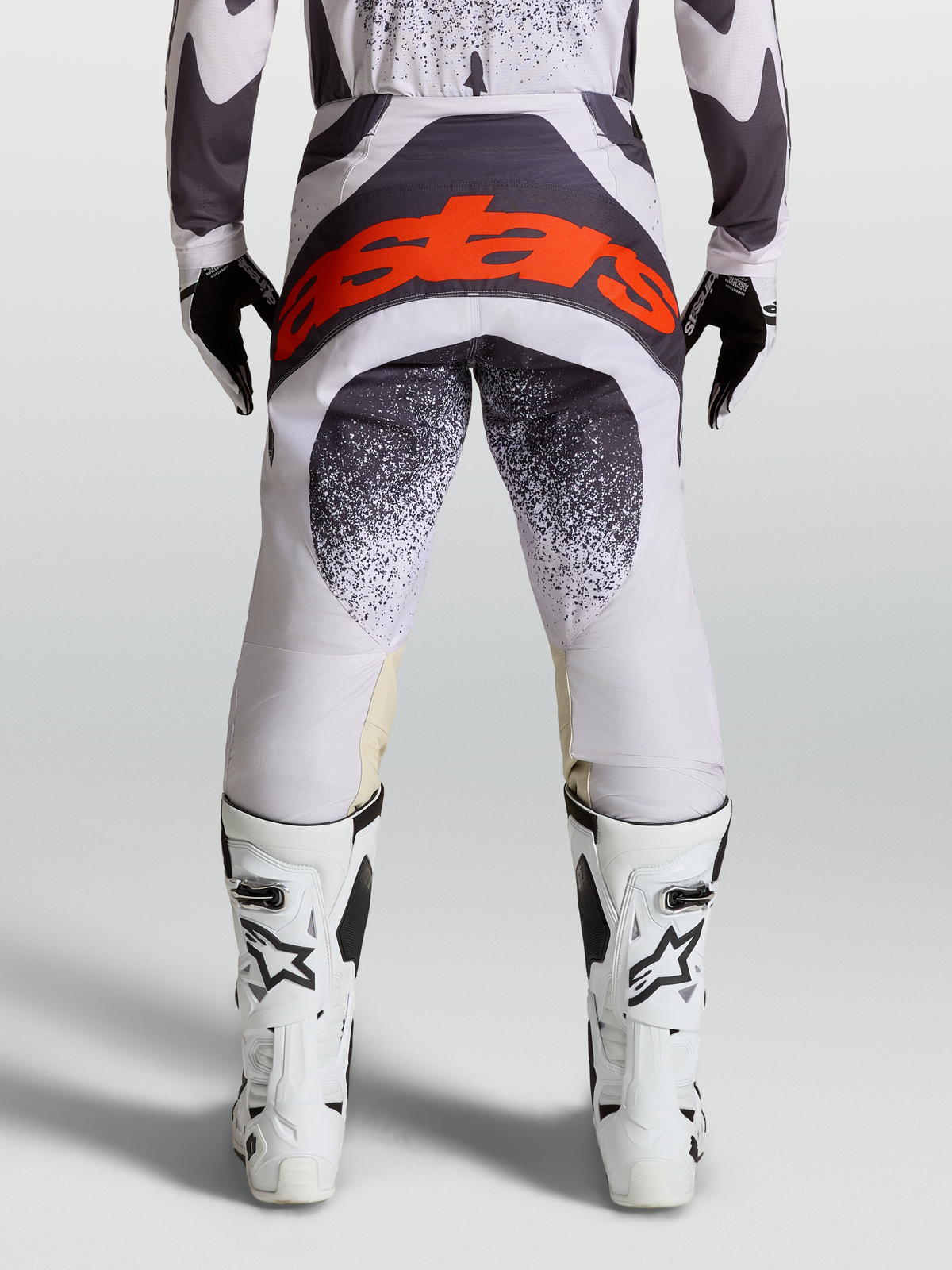 Alpinestars Pantalon Techstar Swan, pantalon de motocross pour homme, lilas clair et gris foncé, vue de dos avec logo Astars rouge fluo, sur un pilote portant des bottes blanches