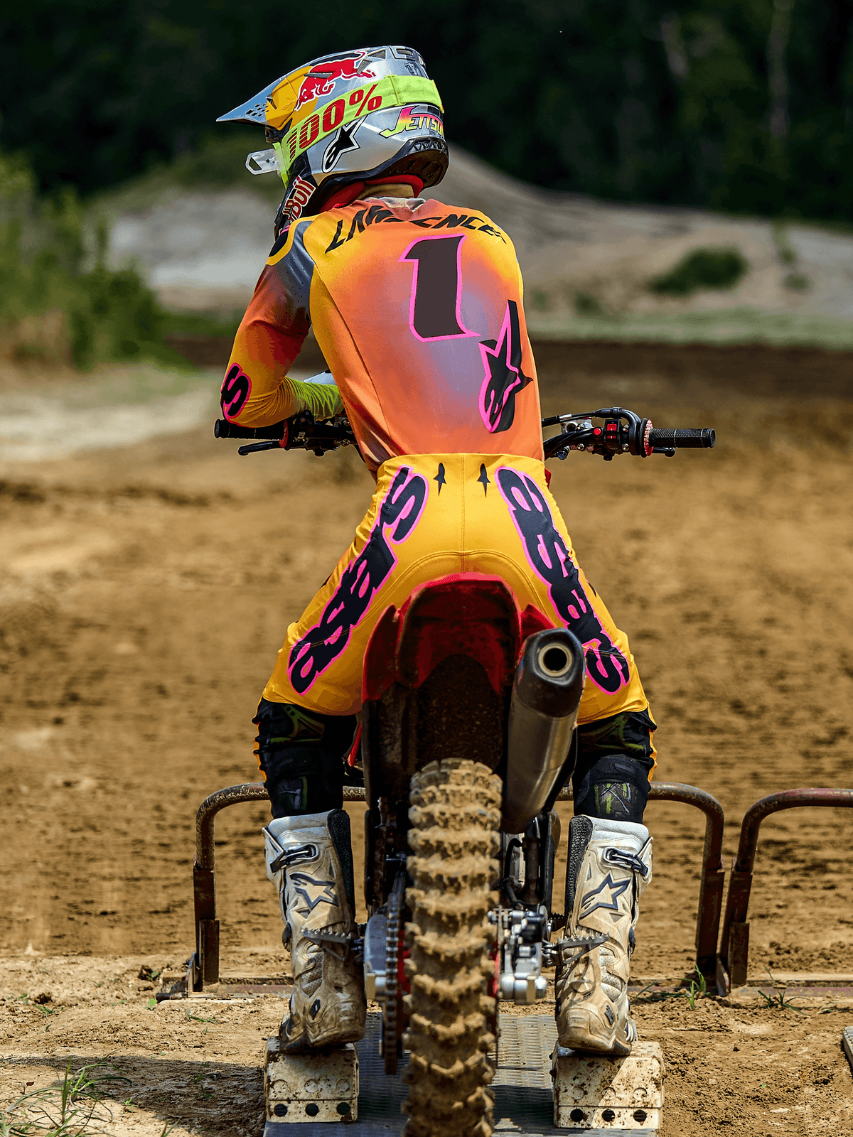 Pilote de motocross de dos portant l'ensemble Alpinestars Supertech Lipan 2025, jaune et rose, maillot avec le numéro 1 et 'LAWRENCE' au dos, pantalon jaune avec marquage 'ASTARS' rose, bottes blanches et casque assorti, assis sur une moto tout-terrain à la grille de départ