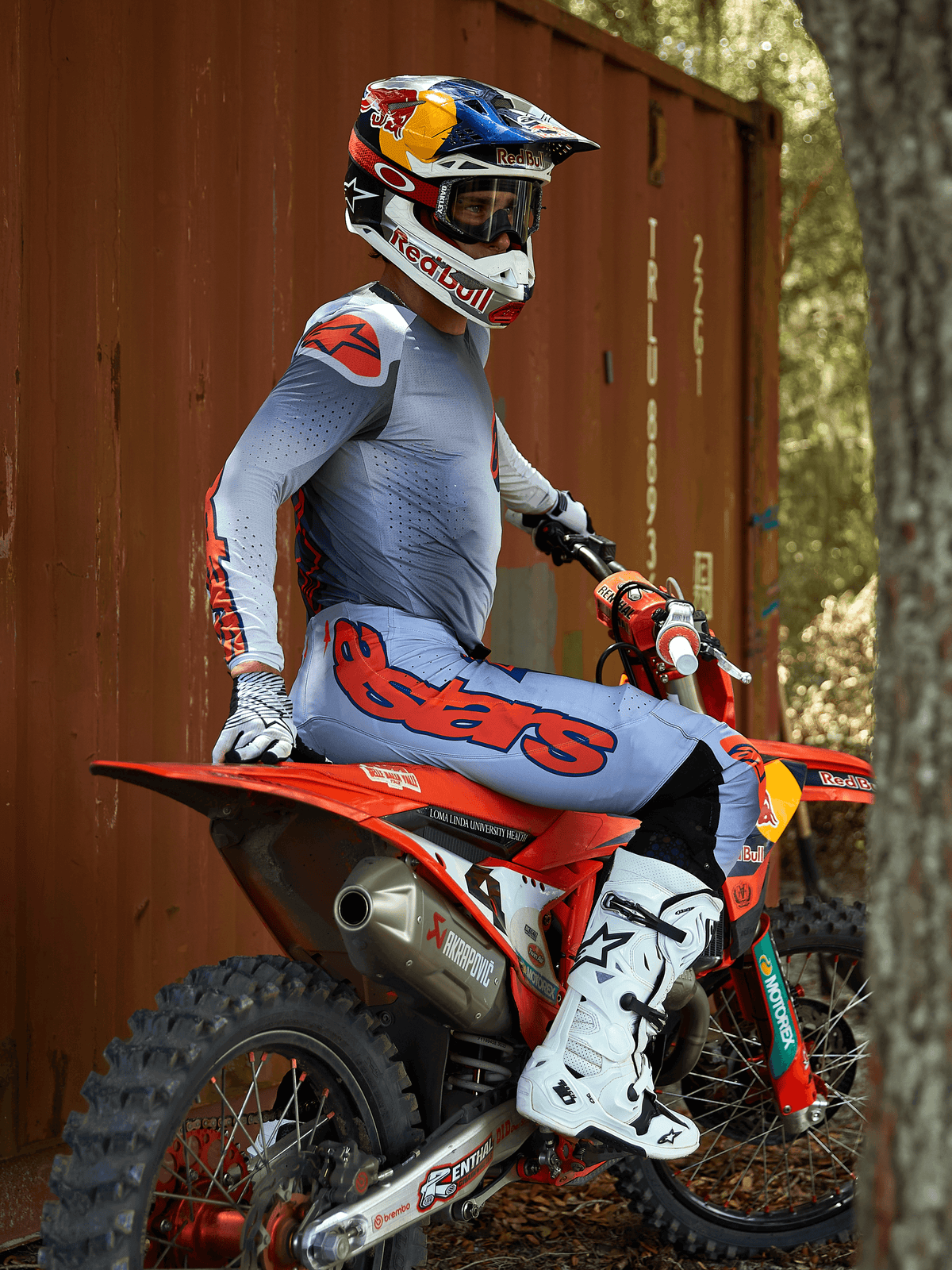Motocross rider sitting on a red dirt bike, wearing the Alpinestars 2025 Supertech Lipan Gear Set in Gray and Orange Fluo, light blue pants with bold orange Astars logo, matching jersey, white motocross boots, and a Red Bull helmet, coordinated professional racing gear