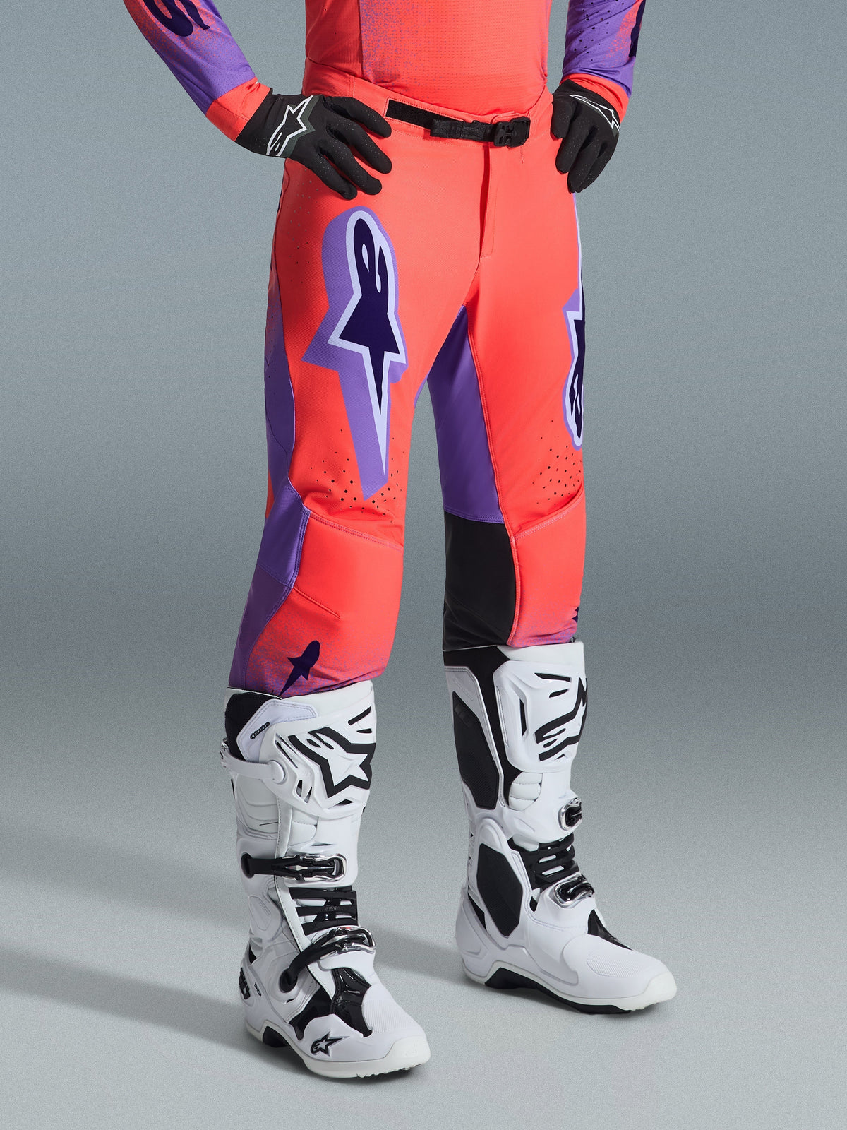 Pantalon Alpinestars Supertech Scenz 2026, pantalon de motocross, corail vif et violet, porté avec des bottes de motocross blanches et noires et des gants noirs, avec un grand logo Alpinestars violet, des panneaux intérieurs aux genoux en cuir noir et une coupe athlétique de course