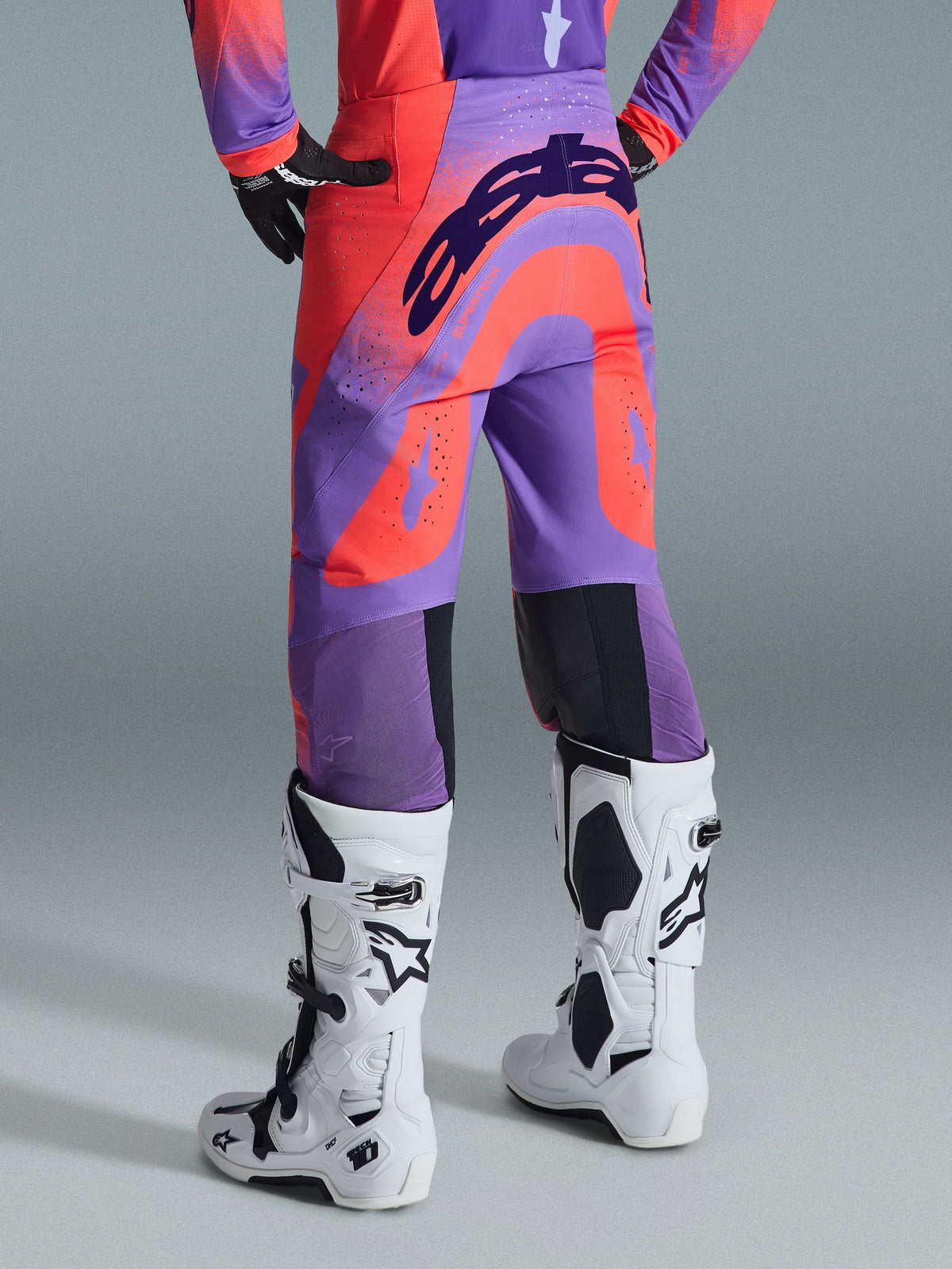 Alpinestars Supertech Scenz 2026, pantalon de motocross, Hot Coral Purple, vue arrière avec marquage audacieux sur l'assise, panneaux corail et violet vifs avec zones perforées pour la circulation de l'air, porté avec des bottes de course blanches et noires
