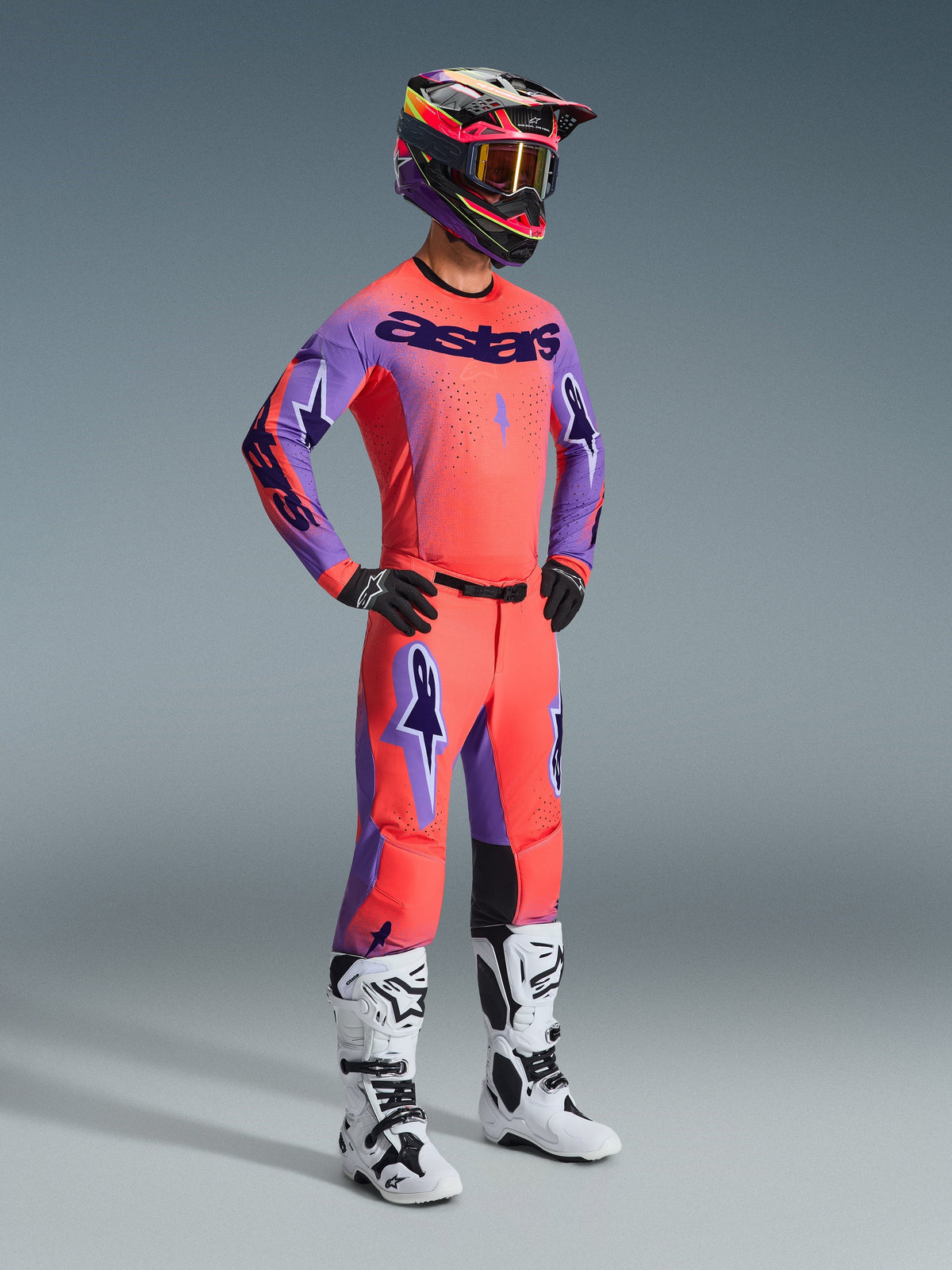 Pilote de motocross portant une tenue de course complète, comprenant le pantalon Alpinestars 2026 Supertech Scenz en Hot Coral Purple, un maillot assorti corail et violet, un casque noir et rose et des bottes blanches et noires, équipement de course tout-terrain professionnel.