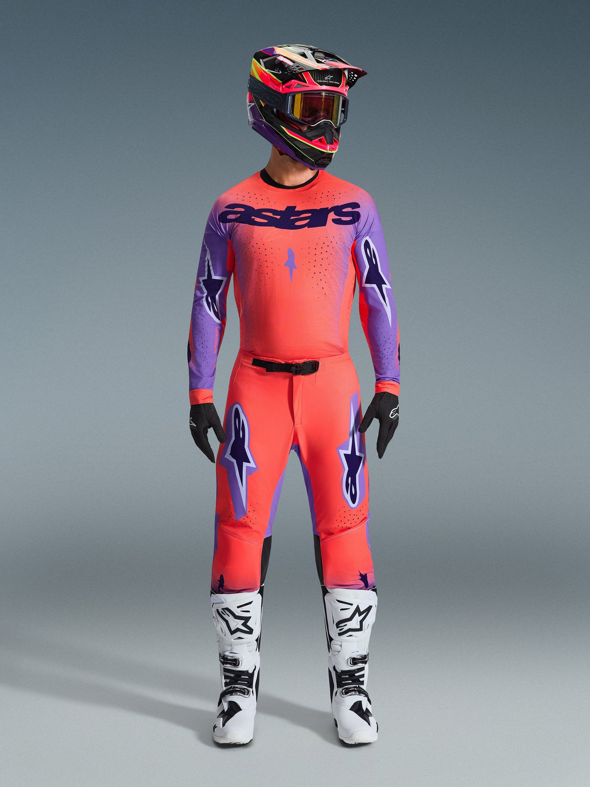 Pantalon Alpinestars Supertech Scenz 2026, pantalon de motocross, corail vif et violet, porté comme une tenue complète avec maillot et casque assortis, bottes blanches et noires, avec logos violets et coupe athlétique de course.