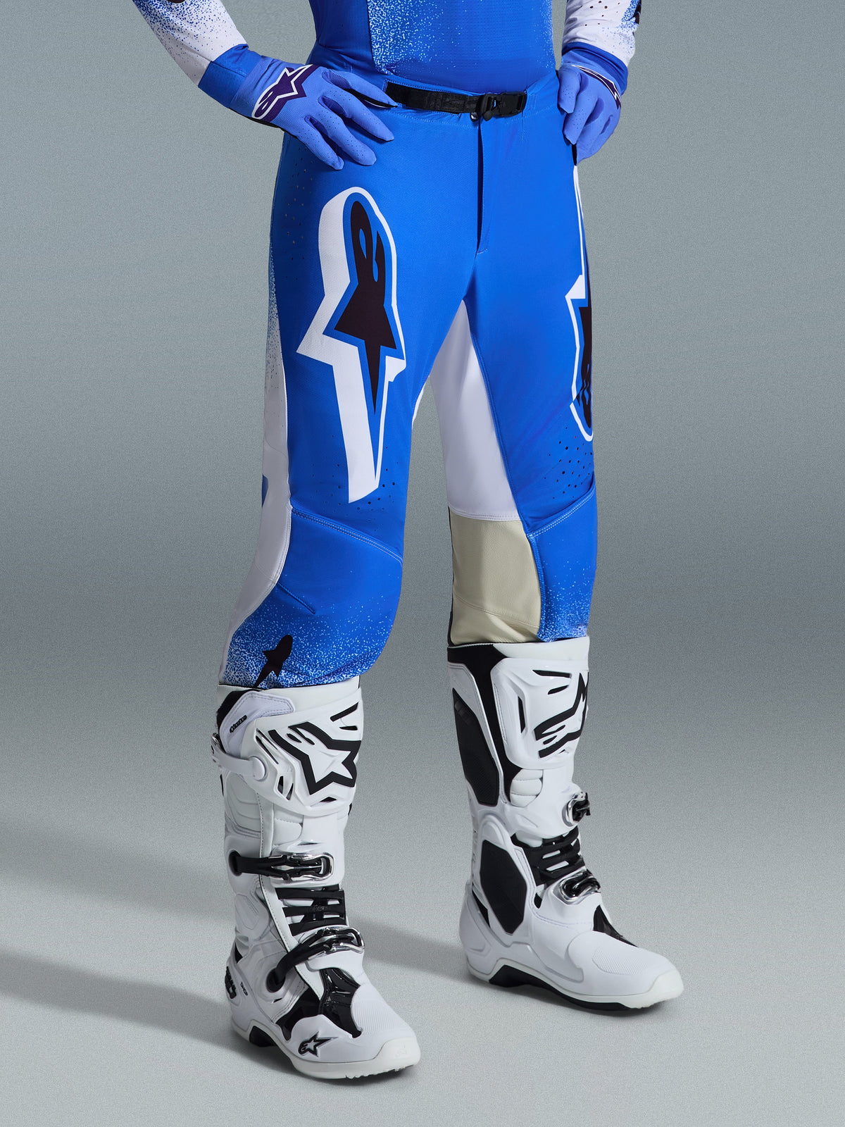 Pantalon Alpinestars Supertech Scenz 2026, pantalon de motocross, bleu UCLA et blanc, porté avec des bottes blanches et noires et des gants bleus, doté de grands logos graphiques sur les cuisses et de panneaux blancs, coupe athlétique conçue pour la compétition de motocross