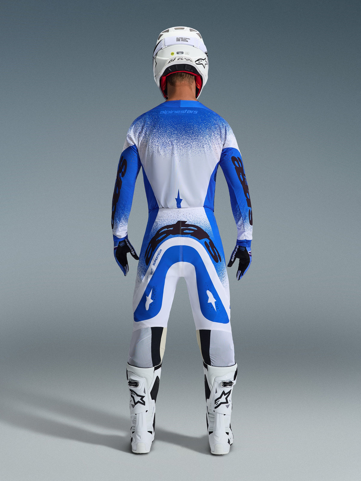 Pilote de motocross de dos portant une tenue de course complète, pantalon Alpinestars Supertech Scenz 2026 bleu UCLA et blanc, maillot assorti bleu et blanc avec motif moucheté, casque blanc, bottes blanches et noires, tenue coordonnée pour la course professionnelle