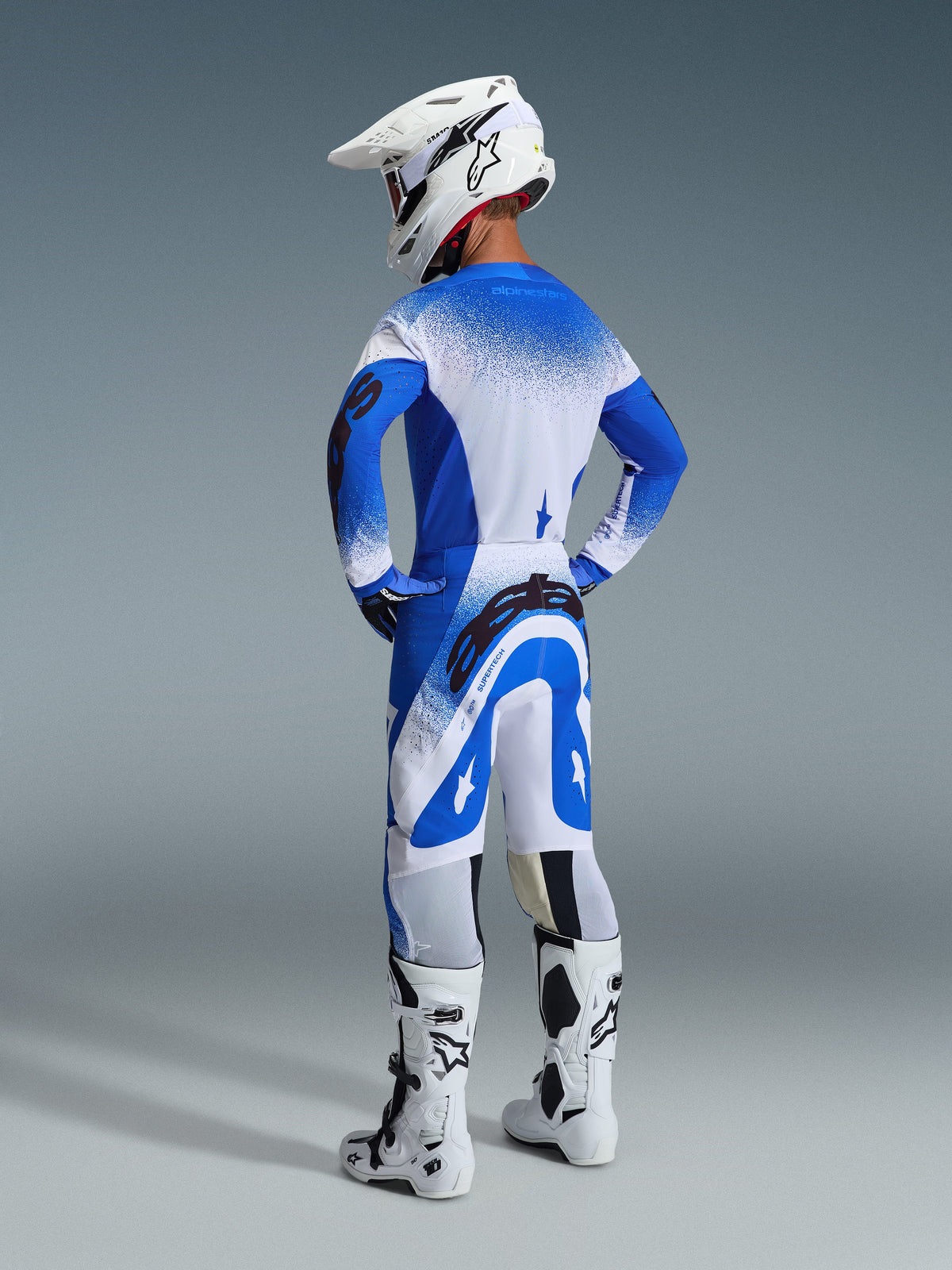Pilote de motocross de dos portant le pantalon et le maillot Supertech Scenz 2026 en Ucla Blue White, casque blanc avec logo Alpinestars, bottes de motocross blanches et noires, ensemble d'équipement de course tout-terrain professionnel