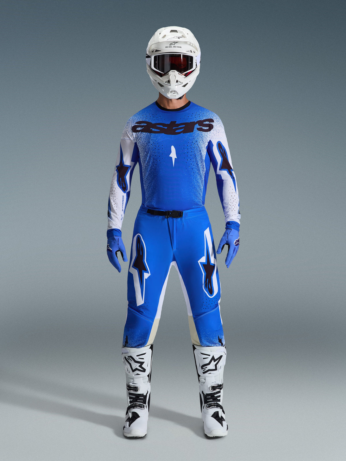 Pilote de motocross portant une tenue de course complète, maillot bleu et blanc avec marquage astars noir, pantalon Supertech Scenz 2026 en Ucla Blue White avec logos étoiles, casque blanc, gants bleus et bottes blanches, tenue de course athlétique coordonnée