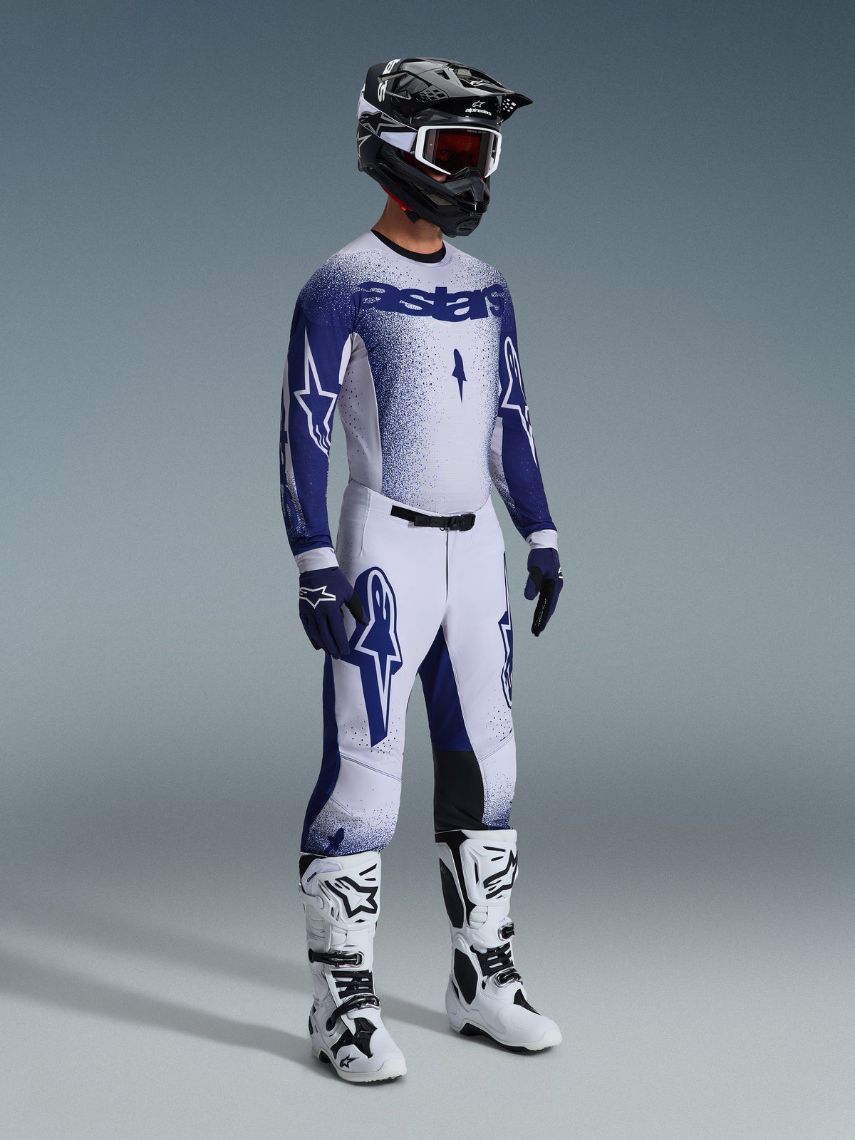 Motocross rider wearing 2026 Supertech Scenz Pants in Light Gray and Dark Blue, matching blue and white jersey with 'astars' branding and spray-paint effect, black helmet with white goggles, white and black motocross boots, coordinated professional off-road racing outfit.