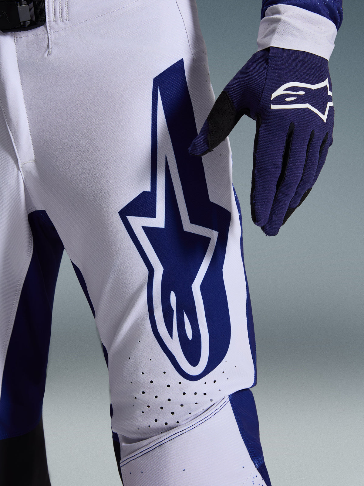 Alpinestars 2026 Supertech Scenz Pants, Motocross Pants, Light Gray and Dark Blue, close-up view of the thigh showing a large dark blue Alpinestars logo, perforated ventilation zones, and stretch rip-stop fabric, worn with a matching dark blue motocross glove