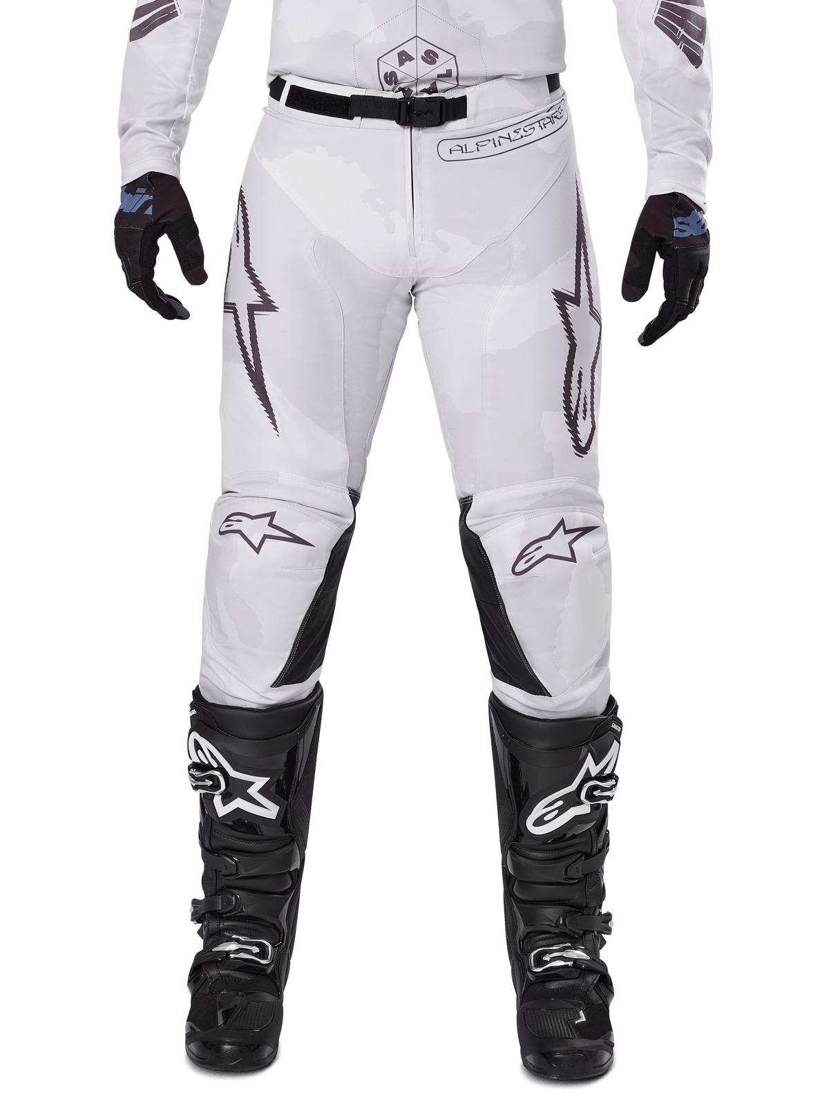 Pantalon Alpinestars 2025 Racer Hollow, pantalon de motocross, camouflage brun clair, porté avec des gants noirs et des bottes de motocross, doté d'un motif camouflage subtil avec des graphismes noirs et les logos Astars, conception ergonomique pour une conduite tout-terrain performante