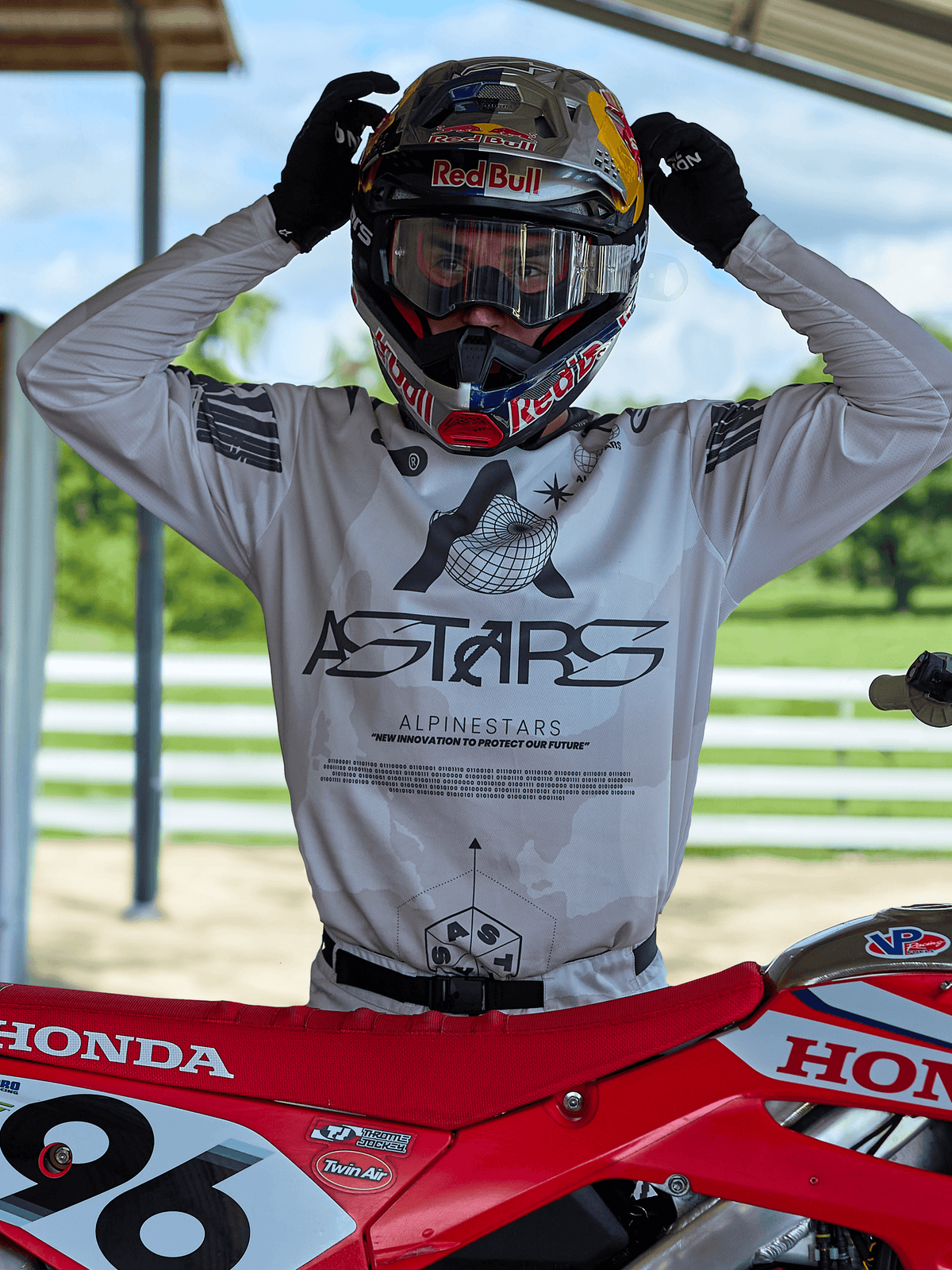 Pilote de motocross portant l'équipement Alpinestars 2025 Racer Hollow, maillot camouflage clair avec marquage ASTARS imposant, ajustant un casque Red Bull muni de lunettes transparentes, debout à côté d'une moto Honda rouge dans un cadre extérieur tout-terrain