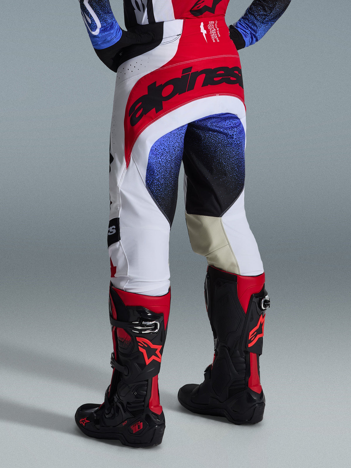 Pantalon Alpinestars Techstar Knif 2026, pantalon de motocross, noir, blanc et rouge avec accents bleus, vue arrière montrant le logo distinctif à la taille et les empiècements intérieurs aux genoux en cuir beige, porté avec des bottes noires et rouges, équipement de course haute performance pour la compétition tout-terrain