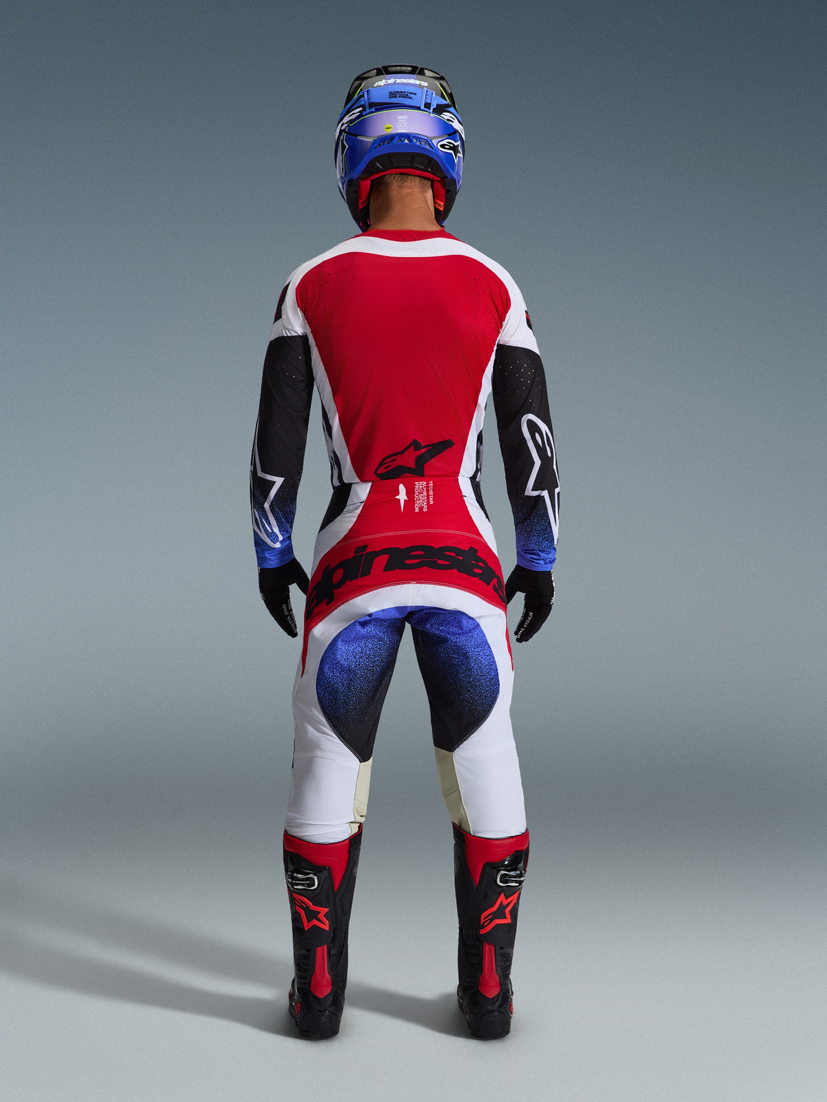 Alpinestars Pantalon Techstar Knif 2026, pantalon de motocross, noir, blanc, rouge et bleu, vue arrière d'un pilote en tenue complète avec maillot assorti, casque bleu et bottes noires et rouges, avec logo Alpinestars distinctif sur l'assise.