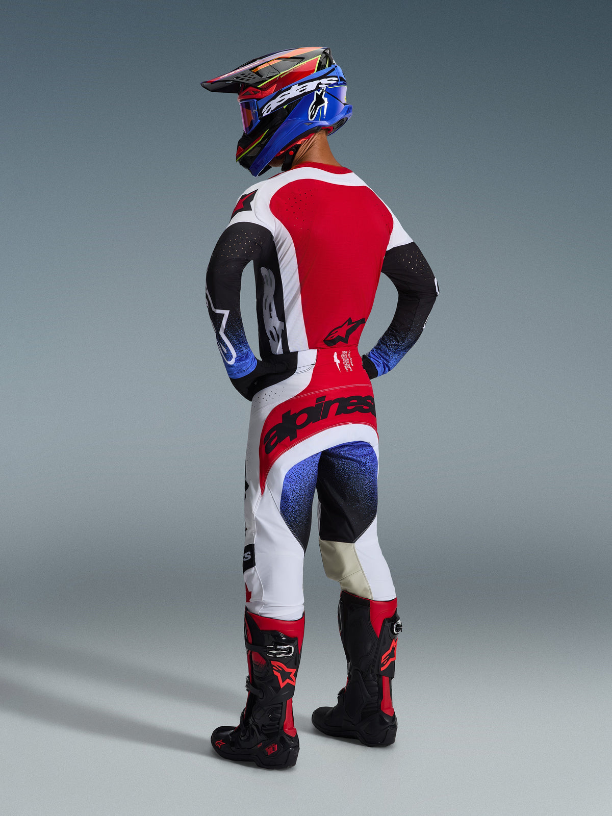 Pantalon Alpinestars Techstar Knif 2026, pantalon de motocross, noir, blanc et rouge avec accents en dégradé bleu, vue arrière d'un pilote portant un équipement de course complet, logo rouge vif sur l'assise, empiècements blancs et noirs, porté avec maillot assorti et bottes Tech 10.