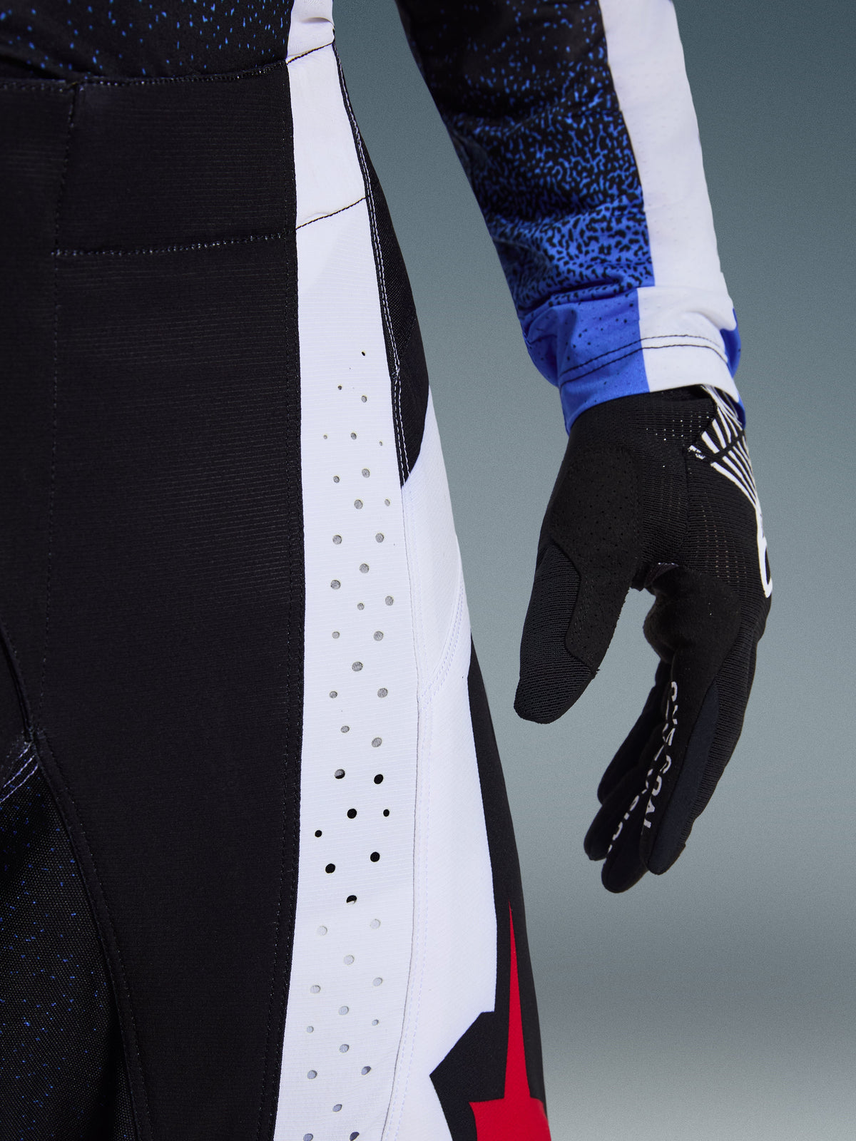Pantalon Alpinestars Techstar Knif 2026, pantalon de motocross, noir, blanc et rouge, gros plan mettant en évidence les perforations découpées au laser sur les panneaux latéraux et le logo étoile rouge, associé à un maillot assorti et un gant de motocross noir