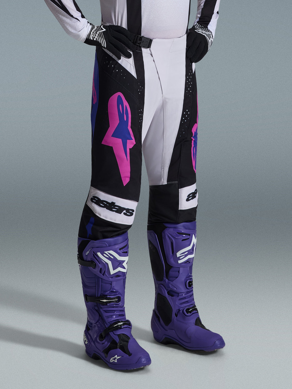 Pantalon Alpinestars Techstar Knif 2026, noir, gris clair et violet, porté avec des bottes de motocross violettes assorties, avec des accents rose vif et le marquage 'astars', équipement de motocross professionnel