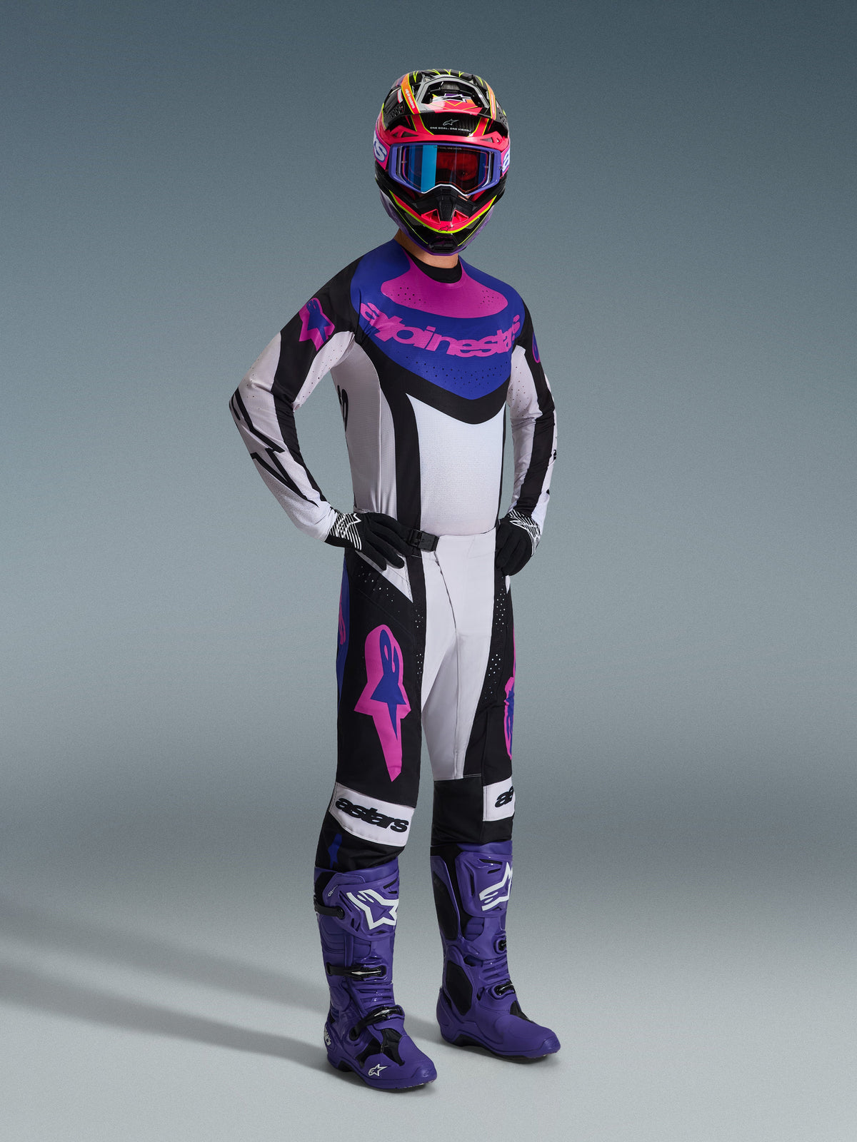 Pilote de motocross portant l'ensemble Alpinestars Techstar Knif 2026, maillot blanc avec accents noirs, bleus et roses, pantalon assorti avec logos Astars roses, casque multicolore avec lunettes, bottes de course violettes, équipement haute performance coordonné pour la compétition