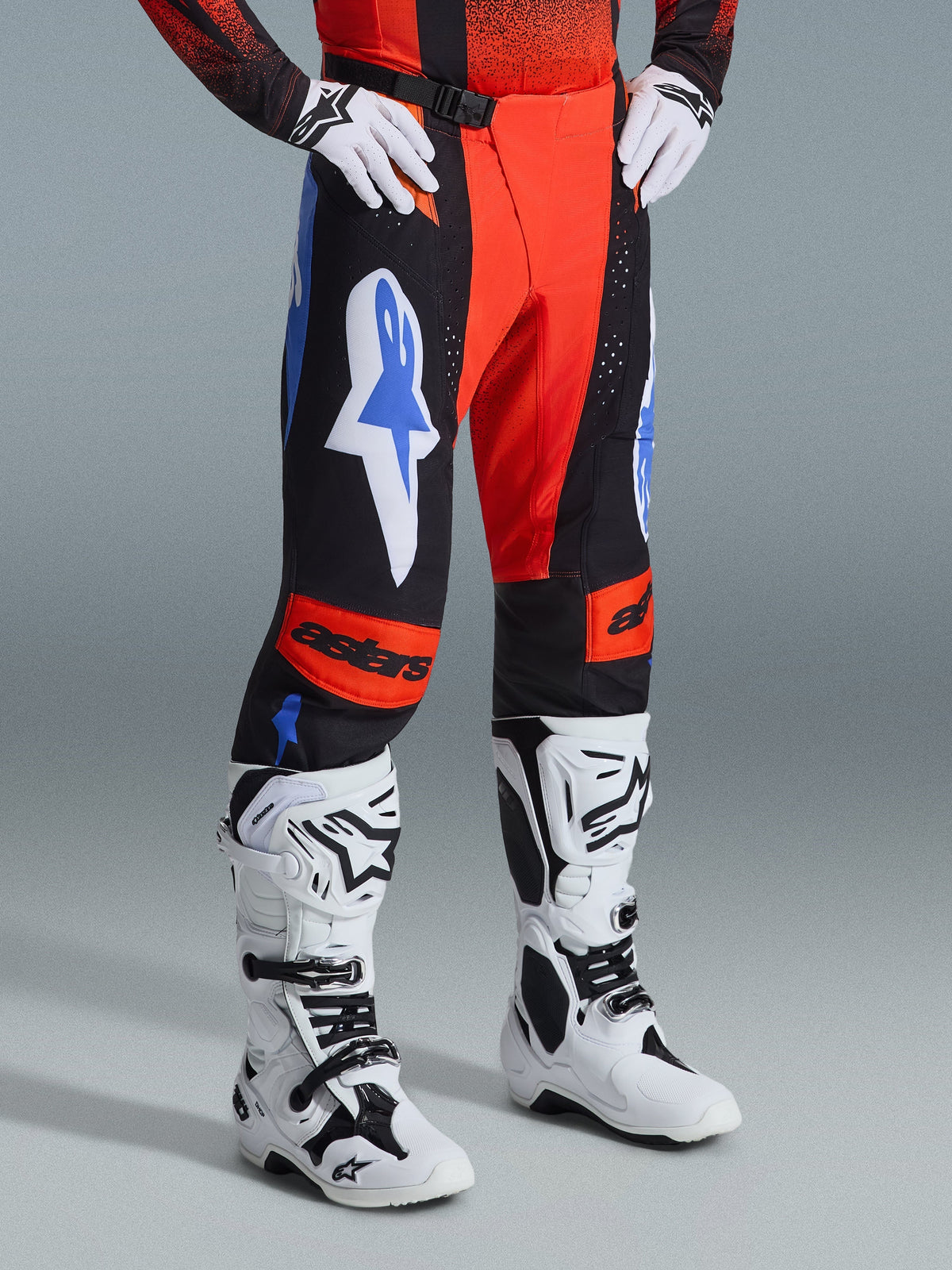 Pantalon Alpinestars Techstar Knif 2026, pantalon de motocross, orange, noir et bleu UCLA, porté avec des gants blancs et des bottes de motocross blanches et noires, doté de logos imposants sur les cuisses et d'empiècements orange aux genoux avec marquage astars.